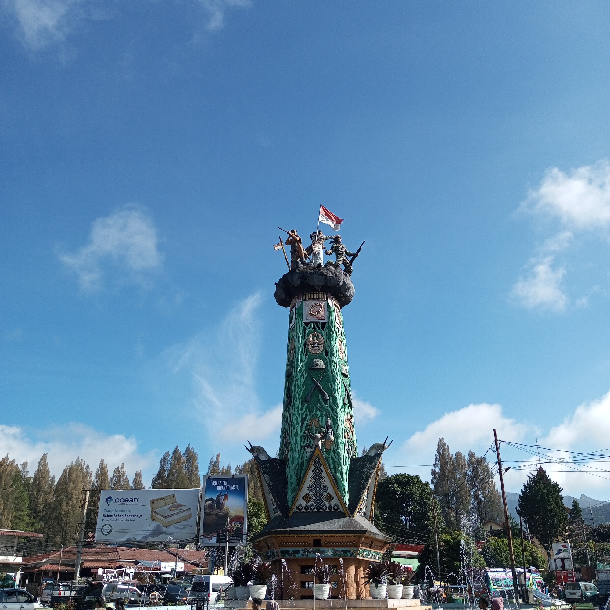 The image shows a monument in the city of Berastagi, Indonesia, under a bright blue sky with scattered clouds. The monument, predominantly green, features sculpted figures at its apex, including individuals holding rifles and an Indonesian flag. The base of the monument is adorned with intricate patterns and decorative elements, with a fountain operating at its lower section, spraying water into the air. Surrounding the monument are trees, buildings, and billboards, one of which advertises "Ocean" mattresses. Vehicles are visible in the foreground, suggesting a public space or roadside location. The overall scene depicts an outdoor environment with a mix of cultural monument, commercial elements, and natural surroundings.