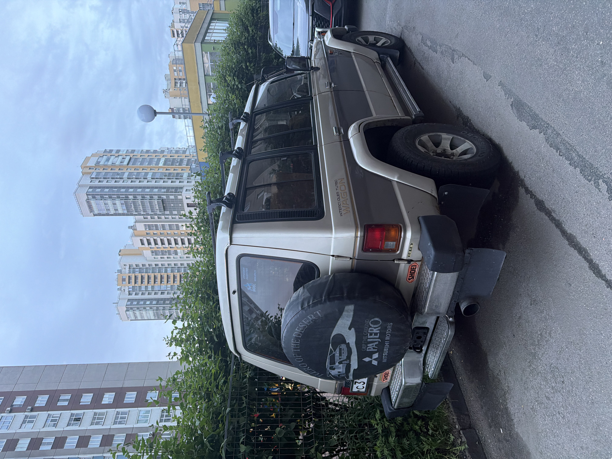 A two-tone silver and brown Mitsubishi Pajero is parked on a paved street in front of a dense green hedge and a chain-link fence. The SUV is positioned at a slight angle to the viewer, facing away from the camera. The rear of the vehicle is prominent, displaying a spare tire cover with graphics that include a stylized depiction of the Pajero, the text "ERO OF THE DESERT," "GRIP THE GROUND," "PAJERO," and "MITSUBISHI MOTORS." License plates are visible, one with "C3" and the other with Cyrillic characters. Mudflaps are attached to the rear bumper, and a trailer hitch receiver is present. On the rear window, there is a sticker with Cyrillic text and a logo. The vehicle has roof racks. In the background, tall residential buildings with multiple stories and balconies rise against a cloudy sky. To the right of the Pajero, the front of a dark-colored SUV is partially visible. The ground is asphalt with some wet patches.