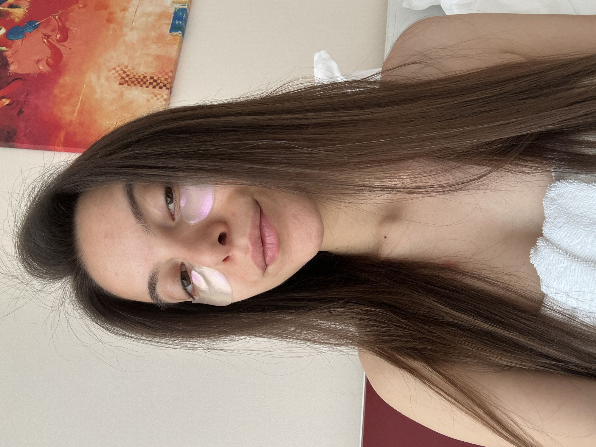 A close-up, front-facing portrait of a young woman with long, dark brown hair, wearing iridescent under-eye patches and wrapped in a white towel. She has light brown eyes and a slight smile. Her hair is slightly messy, framing her face. In the background, a colorful abstract painting with red and orange tones is visible on the wall. To the left of the frame, a section of a dark red headboard is visible, suggesting the setting is a bedroom. The overall ambiance is casual and relaxed, indicative of a personal moment, possibly in a hotel room in Sveti Vlas, Bulgaria, given the context.