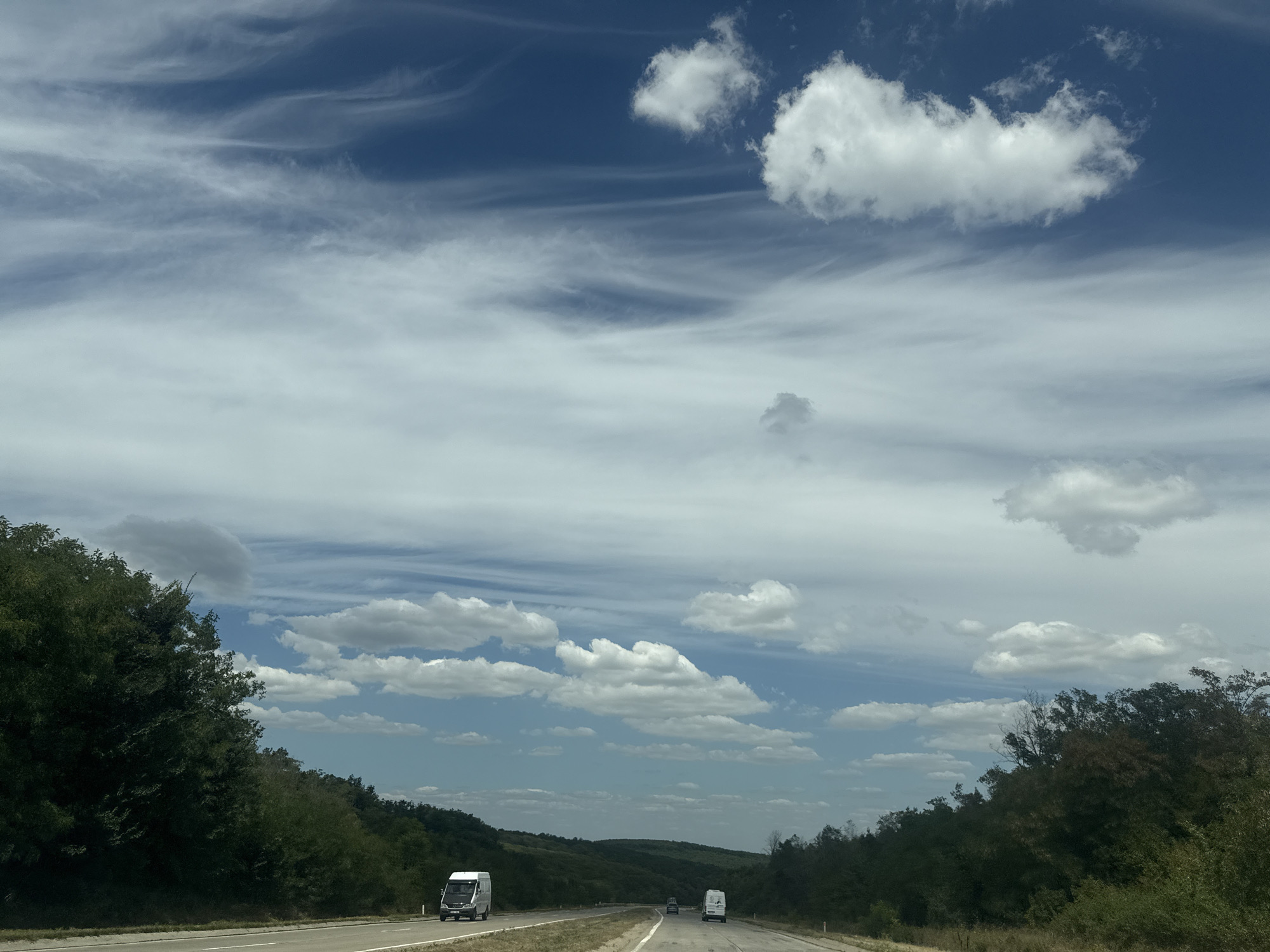 The image shows a view from a vehicle traveling on a highway, likely the M3 highway near the city of Răzeni, Moldova. The road is paved with two lanes in each direction, separated by a white line. On either side of the highway are dense green trees and vegetation, with rolling hills in the distance. The sky is blue with scattered white clouds, some of which are wispy and streaky, indicating wind at higher altitudes. In the foreground, a white van is traveling in the left lane, and further ahead, another white van and a dark-colored vehicle are visible. The overall scene depicts a typical highway drive through a rural, forested landscape under a partly cloudy sky.