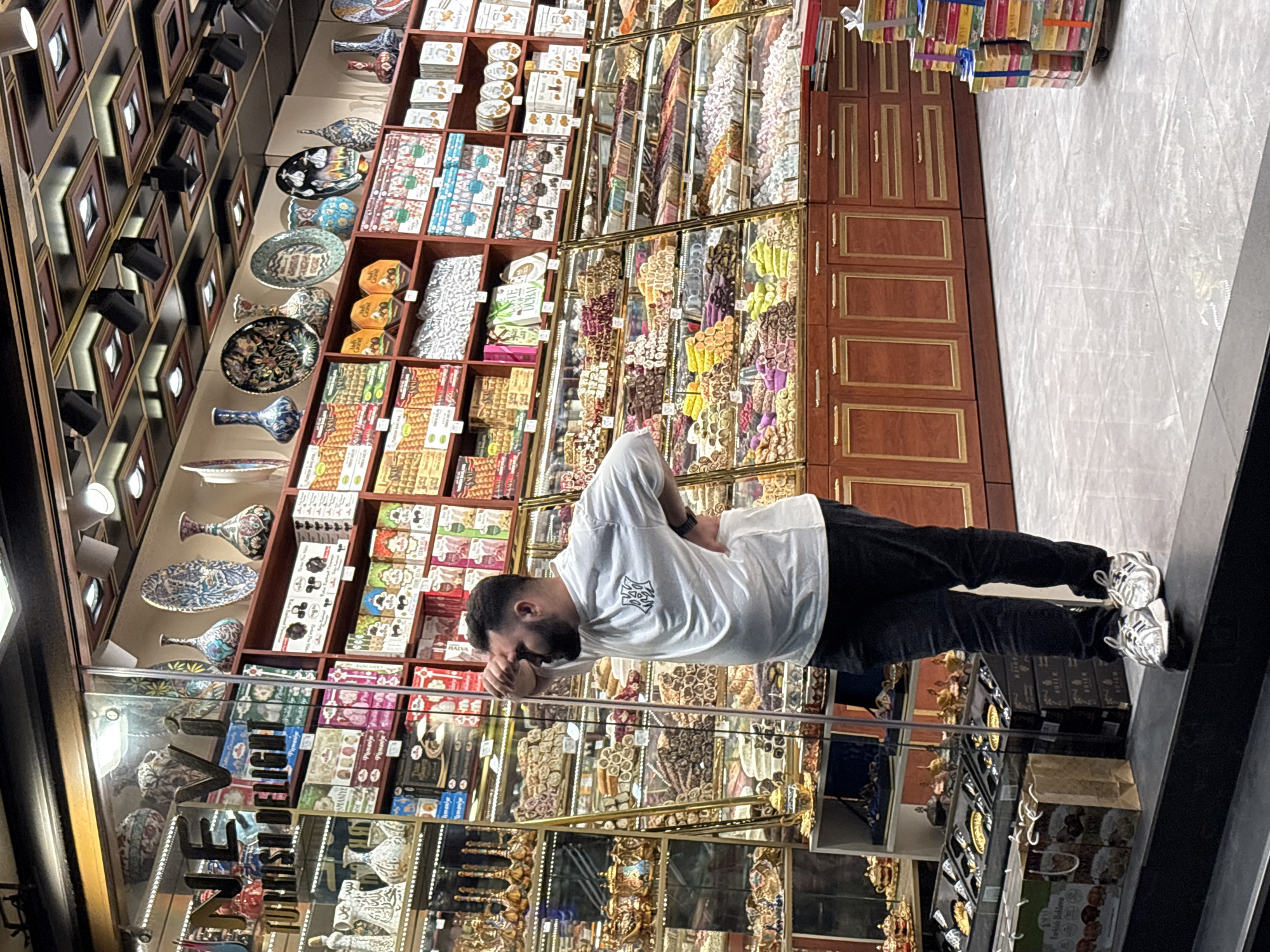 This detailed image captures the interior of a confectionery and gift shop in Beyoğlu, Turkey.
In the foreground, a man with a dark beard and short dark hair stands leaning against a glass display partition or door frame. He is wearing a white short-sleeved t-shirt with a dark, stylized logo on the left chest, dark trousers, and white and grey athletic shoes. His right hand is raised to his forehead, while his left hand rests on his hip, suggesting a thoughtful or resting posture.
The shop interior is brightly lit and features numerous display cases and shelves filled with merchandise. On the left, behind the man and through the glass, shelves showcase various ornate metallic tea sets and other decorative items in gold and silver tones. A partial sign on the glass reads "NEV... TURKISH DELIGHT," indicating the shop's specialty.
Across the mid-ground and background, extensive glass display cases are filled with a wide assortment of colorful Turkish delight (lokum) in various shapes, flavors, and textures. Above these display cases, dark wooden shelves with golden accents hold packaged goods, including boxes of Turkish delight, baklava, and other sweets, some with labels like "Pismaniye" and "Taste of Dubai." The topmost shelves display a collection of decorative ceramic plates and vases featuring intricate patterns and colors, including floral designs and a distinctive garlic motif on some plates.
The shop's ceiling is dark and paneled, adorned with square recessed lights and black track lighting fixtures that illuminate the merchandise. The floor is covered with large, light grey polished tiles. To the far right, a stack of multi-colored gift boxes, secured with ribbons, rests on a small dolly or cart. Wooden base cabinets with golden-framed panel doors and drawers are situated below the main display cases. The overall impression is that of a well-stocked, traditional, and inviting retail establishment.