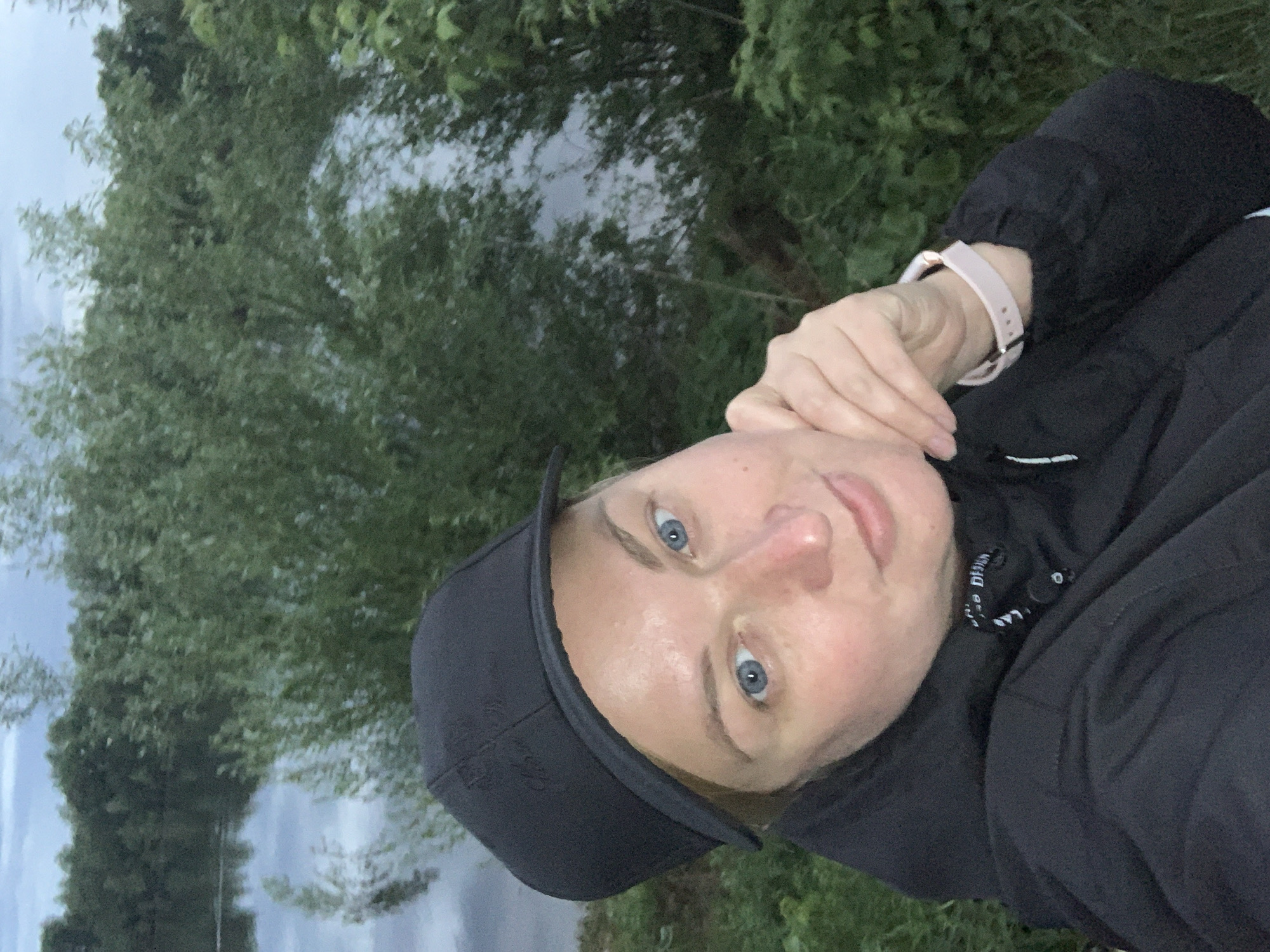 A woman is posing outdoors in a green environment. She is wearing a black cap and a black jacket, and has a pink watch on her left wrist. Her right hand is near her face. In the background, there's greenery, likely trees or bushes, and a body of water, possibly a lake or river reflecting the sky. The location is identified as Chaikovsky, Russia, though this is based on external information and not explicitly visible in the image.