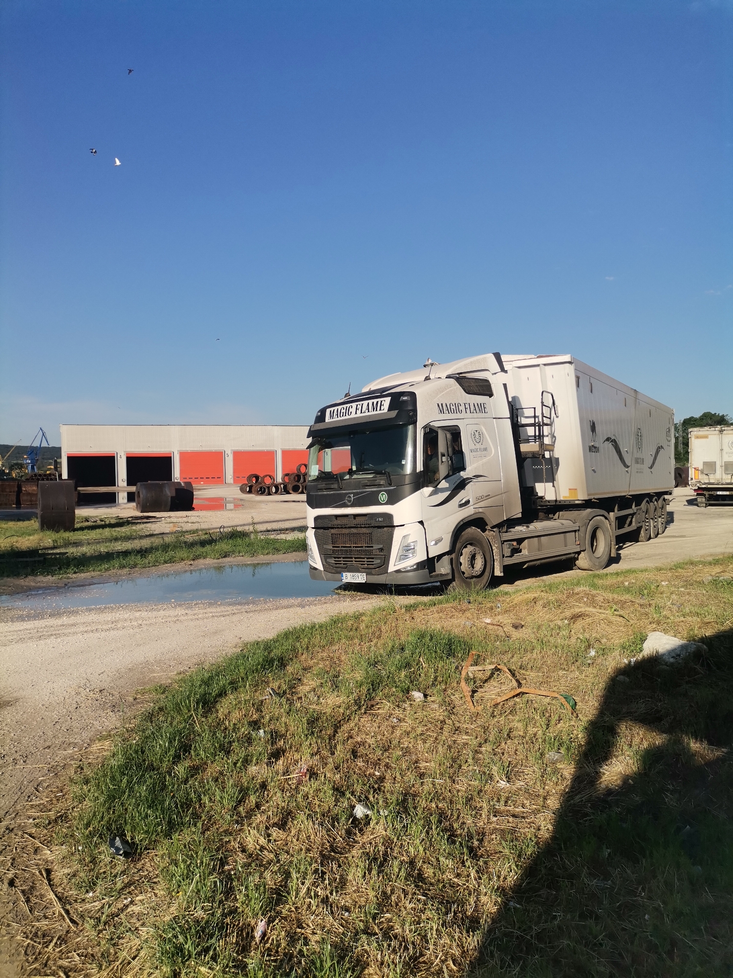 The image depicts a "Magic Flame" truck parked on a dirt road near a puddle in Ezerovo, Bulgaria. Behind the truck is a large white building with five red roll-up doors. A crane is visible in the distance. Old tires are placed in front of the building. The foreground shows a grassy area with some trash. The shadow of the person taking the picture is in the bottom right corner. A few birds are flying in the blue sky. Another trailer is parked nearby.