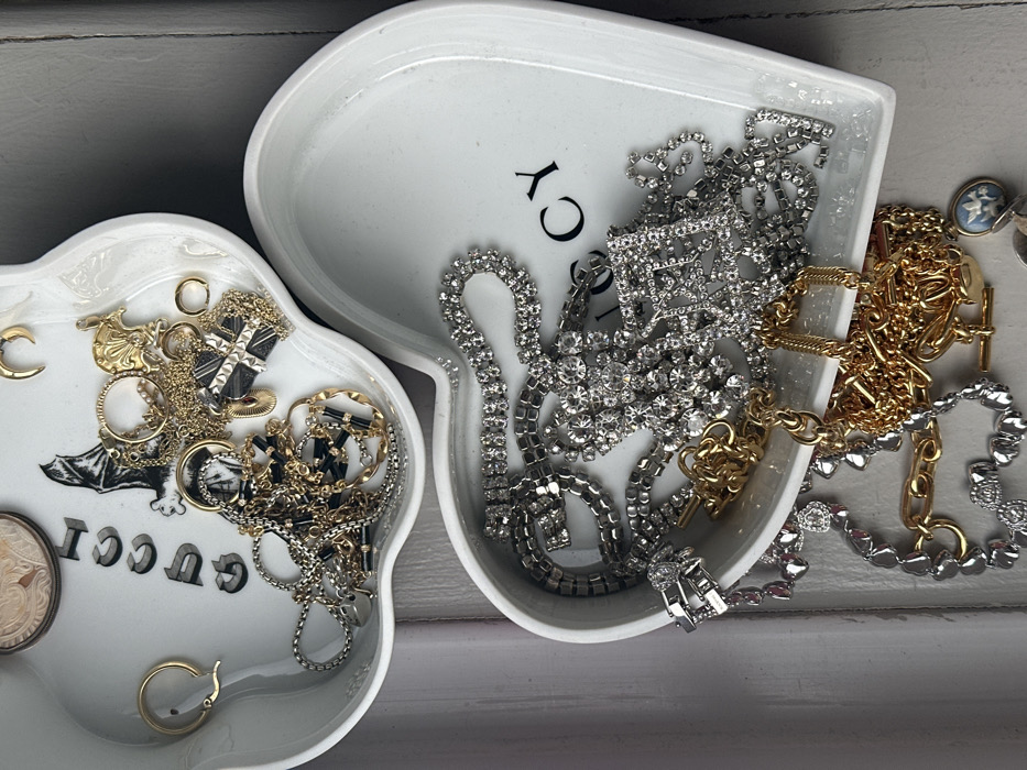 The scene displays two white, heart-shaped ceramic dishes holding an accumulation of jewelry, positioned on a light-colored, vertically paneled surface. The upper dish, partially overlapping the lower one, is marked with black "GUCCI" text and a bat motif. It contains assorted gold-toned and silver-toned items, including a plain gold-toned hoop earring, a gold-toned crescent moon charm, a gold-toned textured disc, multiple gold-toned chains, a silver-toned cross pendant with surface texture, and a silver-toned chain featuring black rectangular segments. A circular object with a raised profile is visible near the "GUCCI" logo. The lower dish, partially visible beneath the upper one, also bears black "GUCCI" text and contains a collection of predominantly silver-toned rhinestone jewelry, such as several strands of clear rhinestones and a large square-shaped pendant or brooch densely set with rhinestones. Additionally, various gold-toned chains are present, one featuring a T-bar clasp, and a small oval pendant adorned with a light blue and white design. No individuals are present, and no active interactions or actions are depicted. The background suggests an indoor environment.