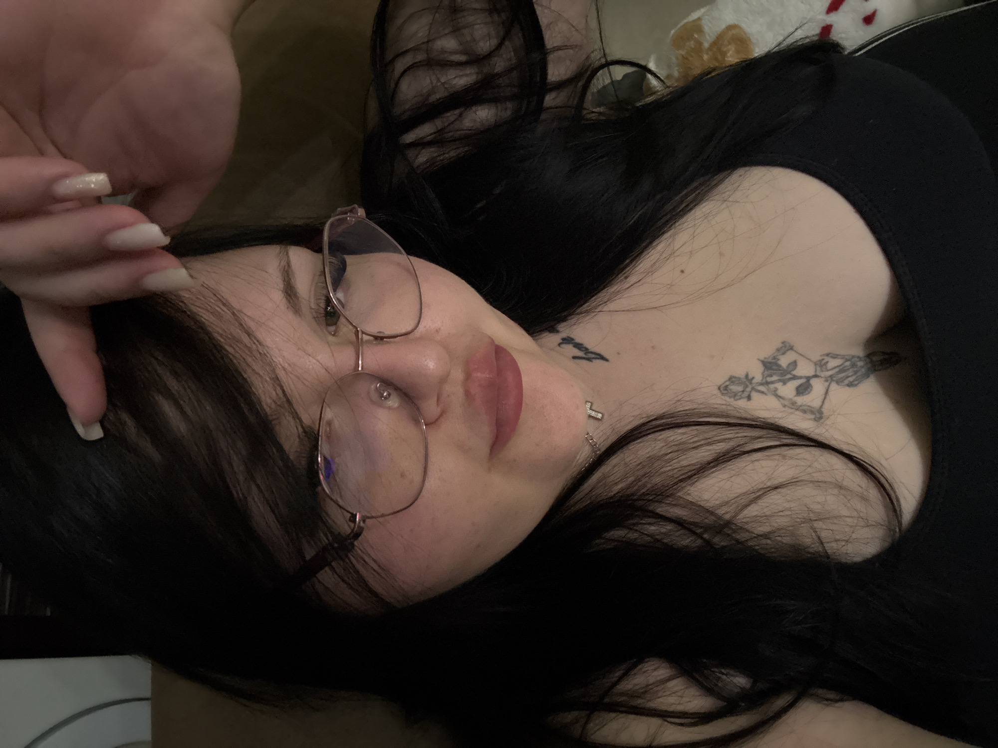 A young woman lies on her back. She has dark hair and wears glasses. There are tattoos on her collarbone and chest. She wears a black top. The background is soft and indistinct. The location is not discernible and cannot be identified as Sofia, Bulgaria based on the available information.