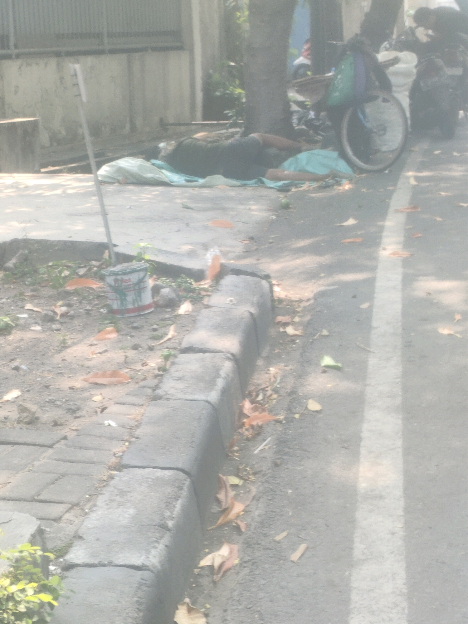A person lies on the ground covered by a light blue sheet, appearing to be asleep or deceased, on a sidewalk next to a tree. A bicycle with a green bag and another object, possibly a motorcycle, are parked nearby. A white and red painted bucket sits on the ground to the left, next to a light pole. The scene appears to be on a street in Surabaya, Indonesia, as indicated by contextual information. The ground is paved with concrete and asphalt, with scattered leaves and debris. Sunlight filters through the trees, casting shadows on the ground.