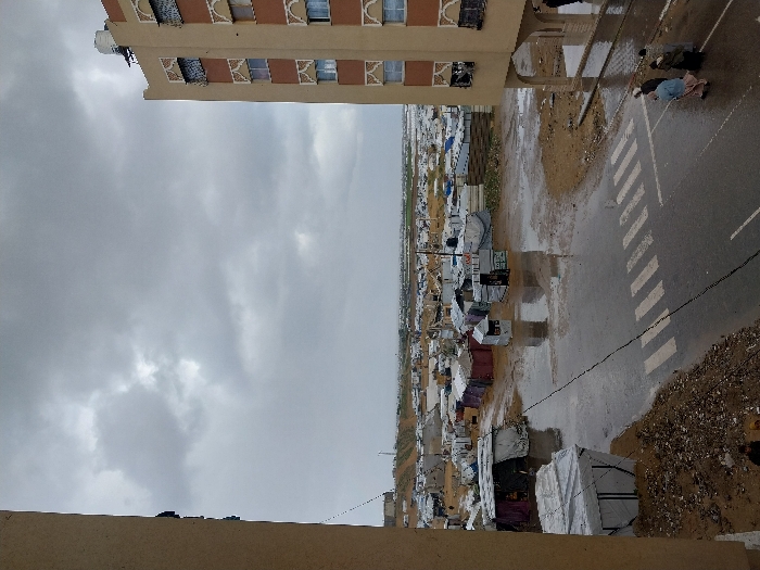 The image presents an elevated view of a temporary settlement in Khan Yunis, Palestinian Territory, under an overcast sky. In the foreground, the left edge shows a partial, plain beige building wall. On the right, a multi-story, beige building with brown decorative elements around its windows stands, featuring a white water tank on its roof.
Below, a wet asphalt road with a marked white pedestrian crosswalk divides the scene. Puddles and muddy areas are visible on the unpaved ground adjacent to the road. Four individuals, appearing as adults, are walking away from the viewer on the right side of the road, near the base of the multi-story building. They are dressed in light-colored and dark clothing.
The midground and background are dominated by a sprawling, dense collection of makeshift shelters and tents. These structures are constructed from various materials including tarpaulins, fabric sheets, and corrugated metal, displaying colors such as white, blue, grey, and maroon. The ground throughout the settlement appears muddy and unkempt, suggesting recent rainfall. Several small, cube-like structures or kiosks are interspersed among the larger tents near the road. The temporary encampment extends far into the distance, meeting a low, barren horizon under a uniform grey, cloudy sky.