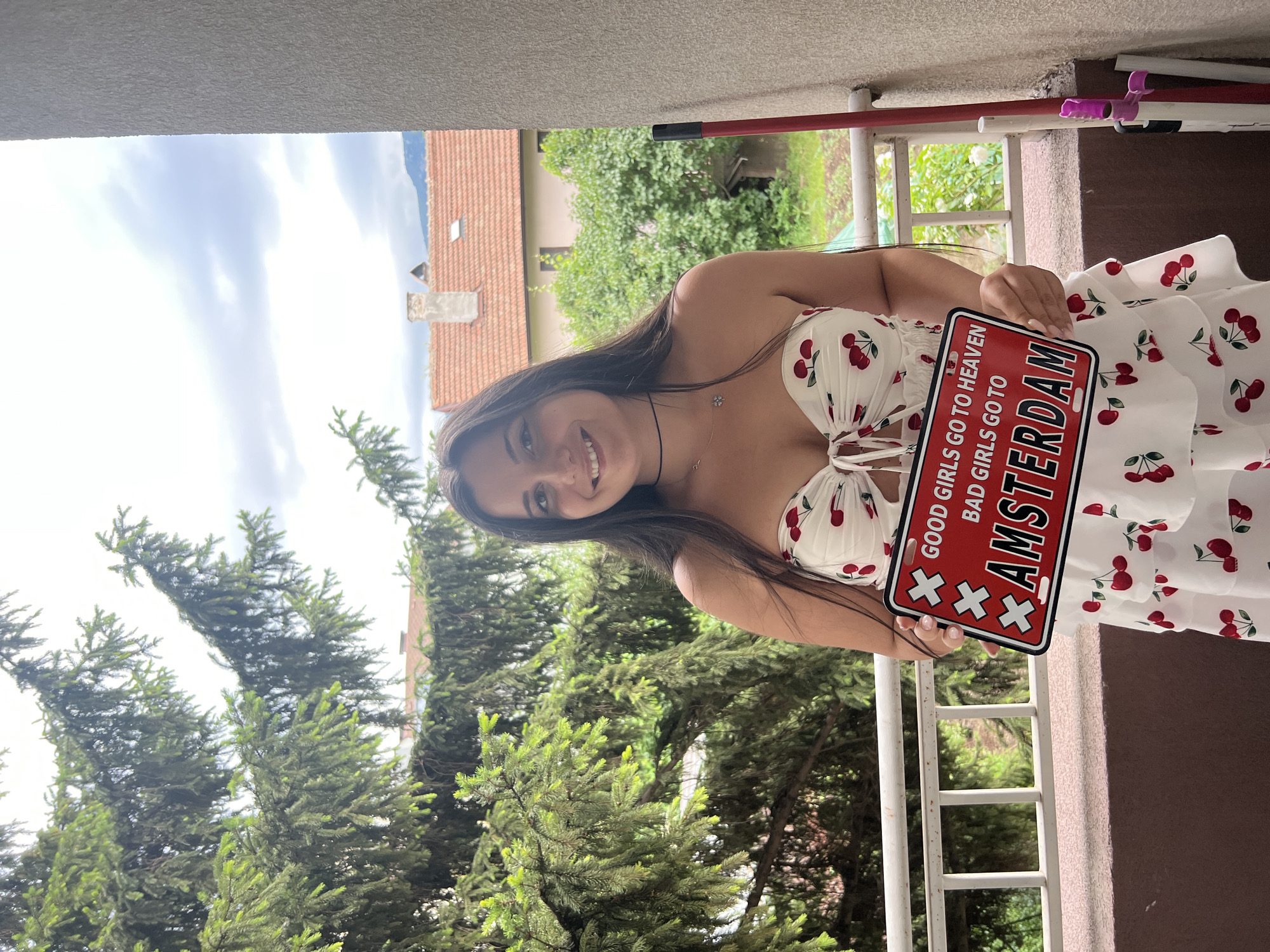 A young woman with long dark hair is standing on a balcony in an outdoor setting, holding a red novelty license plate that reads "GOOD GIRLS GO TO HEAVEN BAD GIRLS GO TO AMSTERDAM." She is wearing a white strapless dress with a cherry pattern and a cutout detail at the bust. The background shows trees, houses with red tile roofs, and a cloudy sky, suggesting a residential area, possibly in Sofia, Bulgaria. The balcony has a white railing, and to the right, a red mop with a purple attachment leans against a textured wall. The overall lighting indicates it is daytime, under an overcast sky.