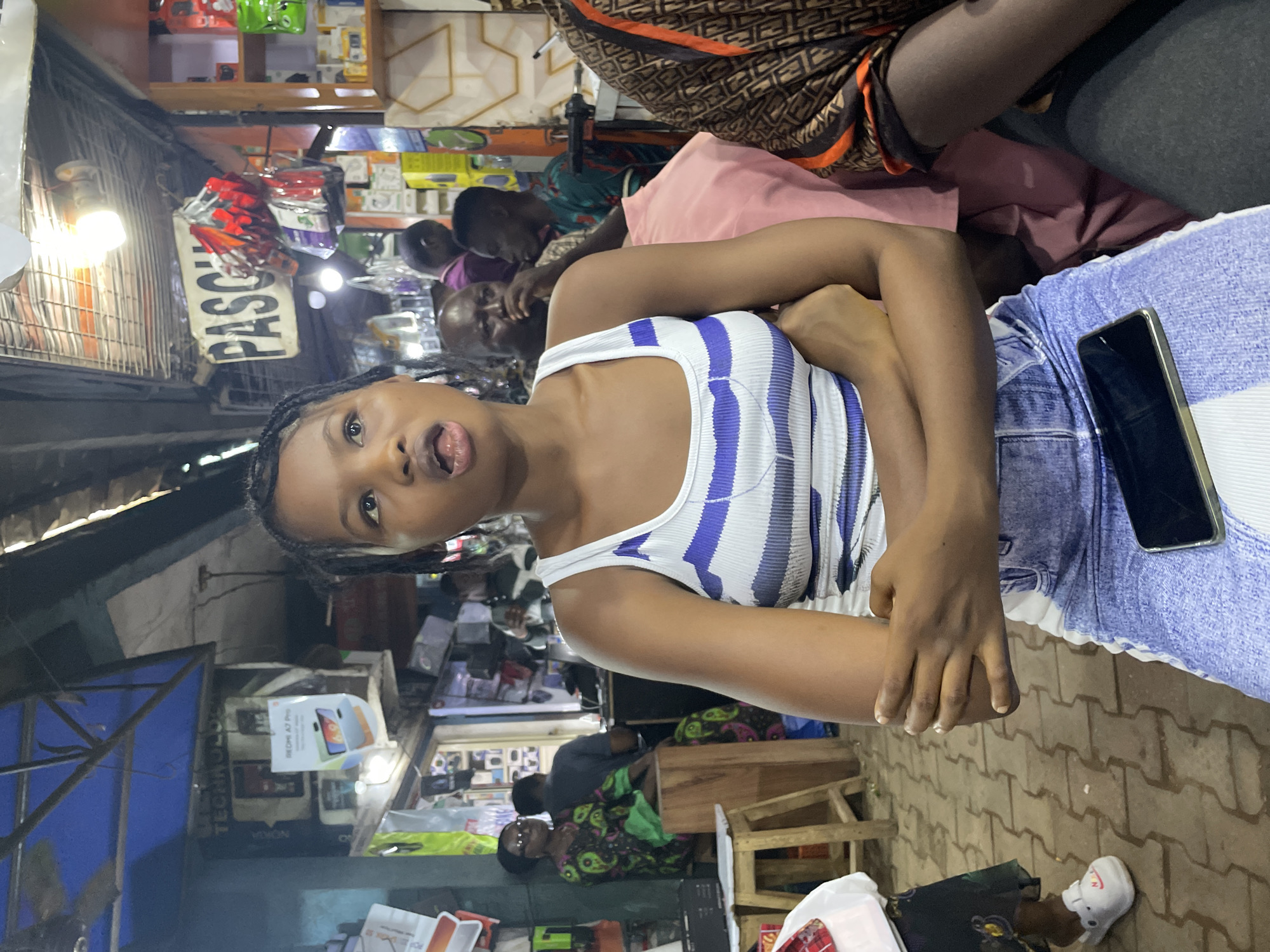 In Nkpor, Nigeria, at a bustling market, a young woman with braided hair, wearing a white and blue striped tank top and denim shorts, sits with her arms crossed. She appears to be looking up and to her right, with her mouth slightly open, as if speaking or reacting to something. A smartphone rests on her lap. Behind her, a stall displays various electronic devices and accessories. In the background, other people are visible, including a woman in a patterned dress and glasses, seated on a wooden stool, and several men. The market setting is indicated by the overhead coverings, exposed wiring, and the arrangement of merchandise.