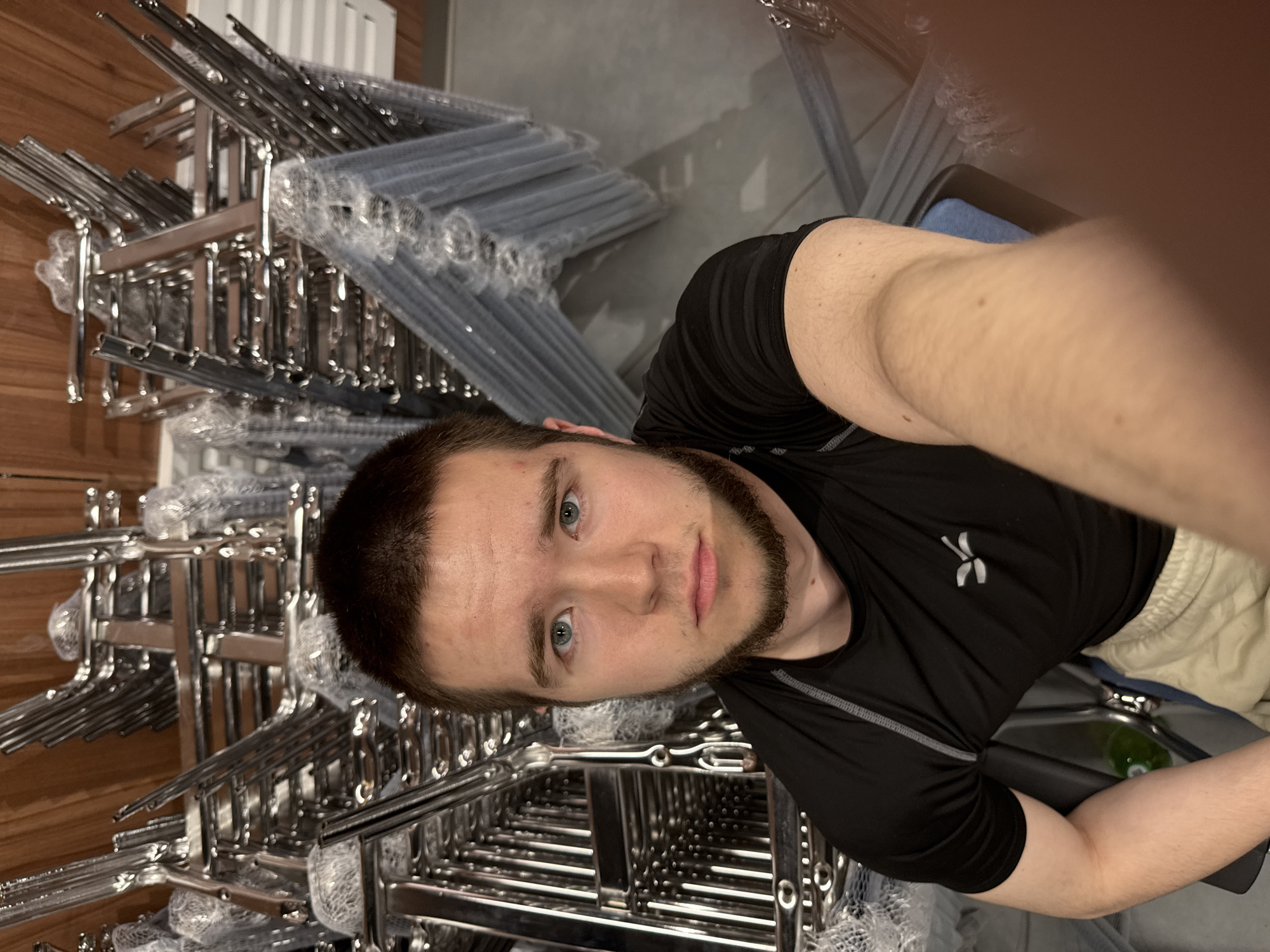 A man is taking a selfie in what appears to be a storage room. He has short brown hair and a beard and is wearing a black shirt. Behind him, there are stacks of metal chairs, many still wrapped in plastic. The environment seems utilitarian, with a mix of wood and gray walls. The location is unknown.