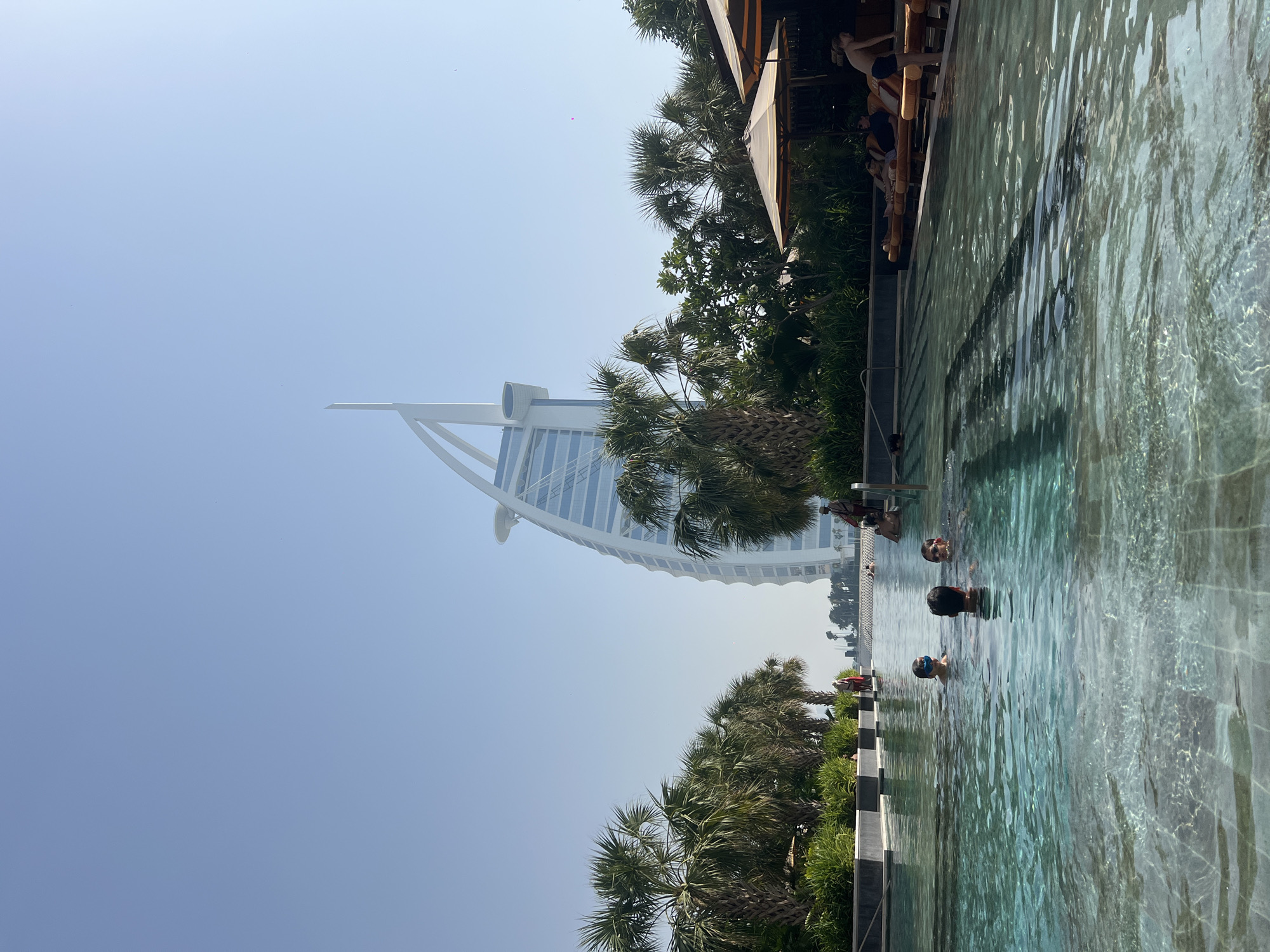 The image shows a view of the Burj Al Arab hotel in Dubai, United Arab Emirates, from a swimming pool. Several people are in the pool. Tropical vegetation lines the pool's edge. The sky is clear and blue.