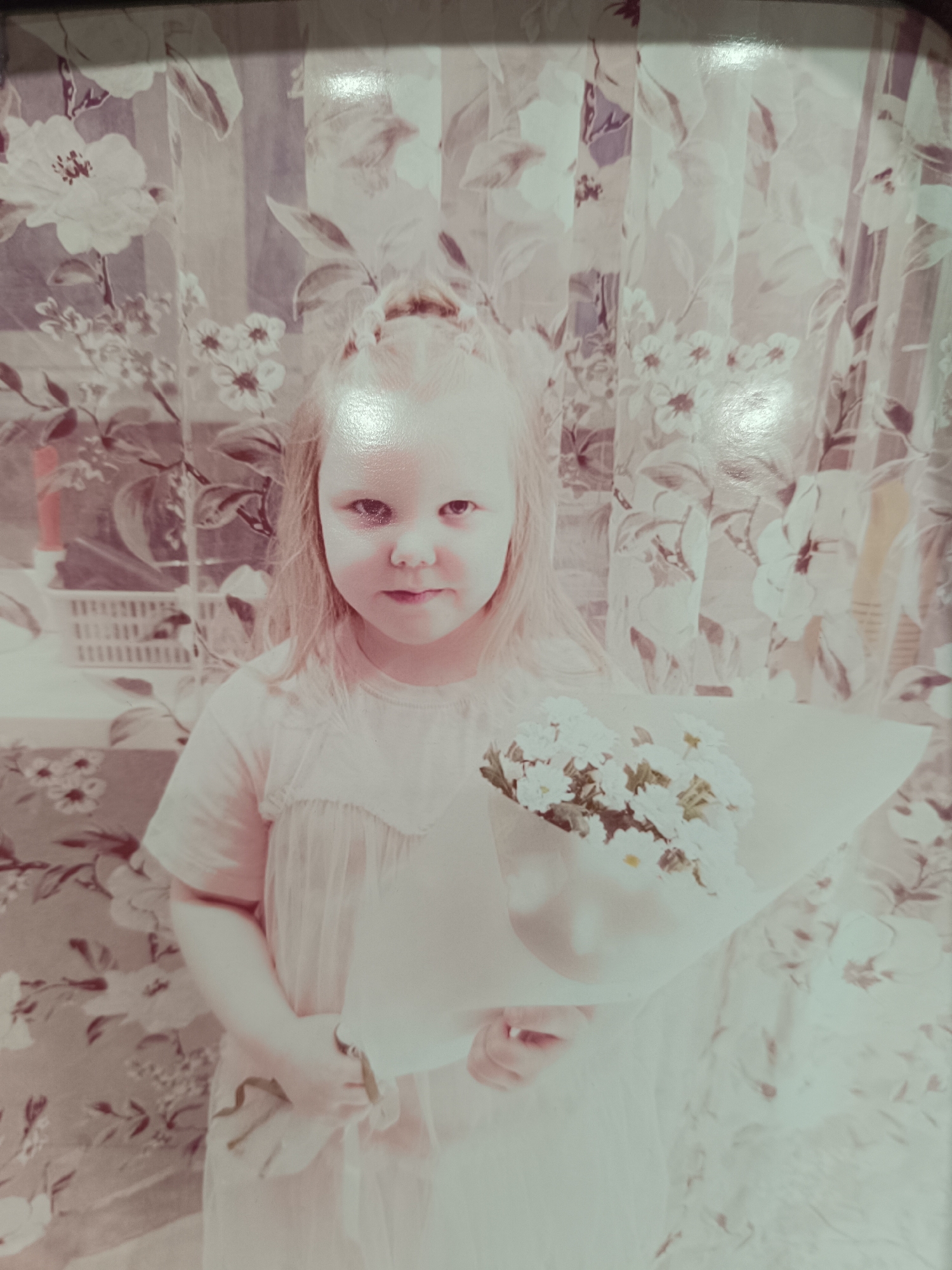 A young girl stands in an indoor environment, possibly a room decorated with floral-patterned wallpaper or curtains. She is holding a bouquet of small, white daisy-like flowers wrapped in paper. She is wearing a light-colored dress. The location is Unknown, Unknown.
