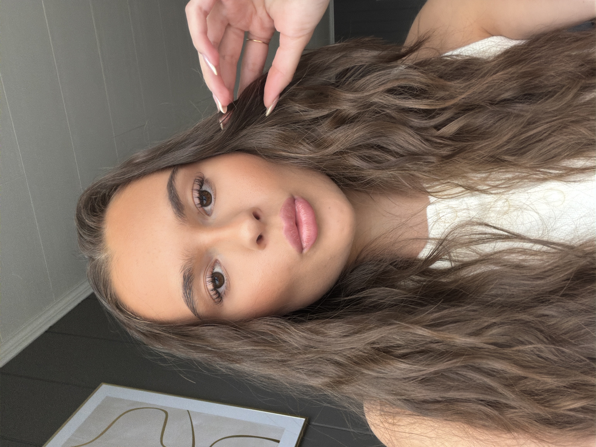 This is a close-up, rotated portrait of a young woman with long, wavy, brown hair. She is looking towards the camera, with her right hand touching her hair near her temple. She is wearing makeup, including mascara and lipstick. She is also wearing a gold ring on her right ring finger. The background is dark, with a lighter-colored wall or partition visible on the left side of the image, and a framed picture on the lower left. There is no visible text in the image. The lighting suggests it is daytime, likely indoors.