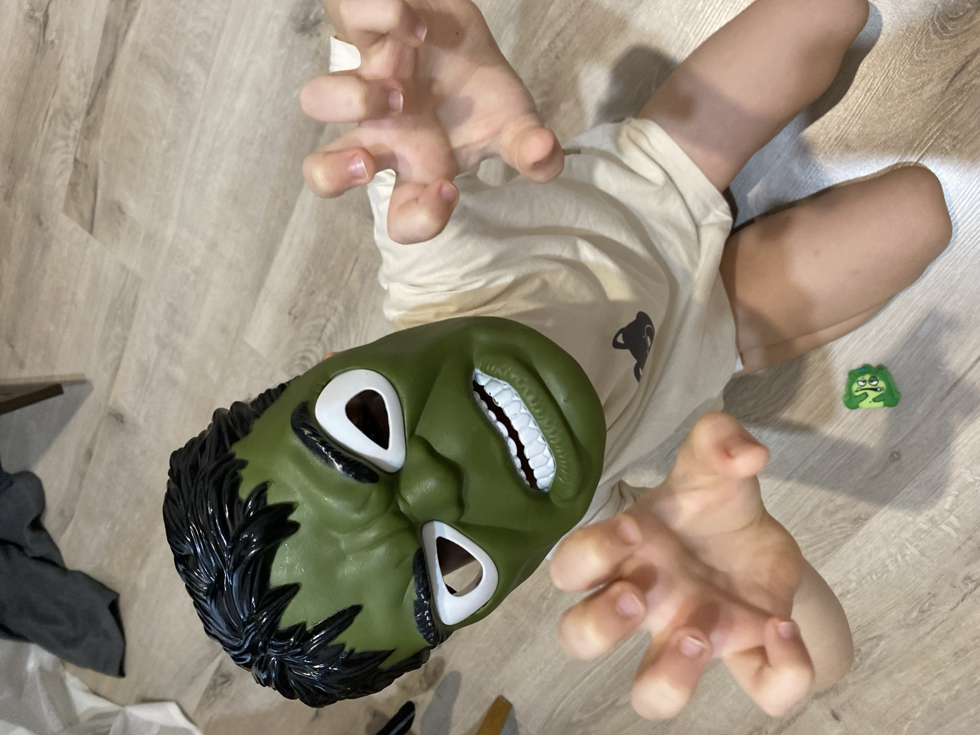 A child wearing a Hulk mask lies on a light-colored, wood-patterned floor. The child is wearing a beige t-shirt. Their arms are raised with fingers outstretched. A small, green, possibly homemade figure rests on the floor near the child's leg. A dark gray garment lies in the background. The location is assumed to be Chaikovsky, Russia based on the provided information.