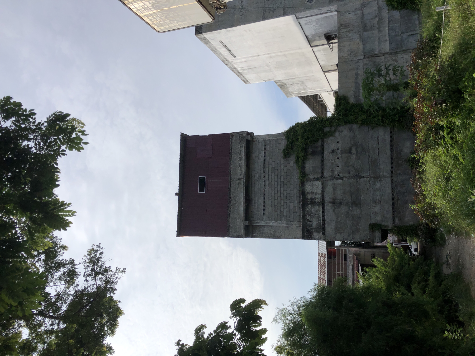 In Lhokseumawe, Indonesia, an overcast sky looms over a multi-story concrete building with a red metal roof. The building is partially covered in climbing vines, and a small, dark rectangular window is visible on the upper section of the structure. To the right, another concrete building with a light-colored facade rises, and a large, reflective, possibly billboard-like structure is visible at the top right of the frame. Lush green foliage, including trees and bushes, surrounds the base of the buildings and encroaches on a dirt path leading towards them. In the distance, another building with a corrugated metal roof is partially obscured by trees. A white car is visible parked on the path.