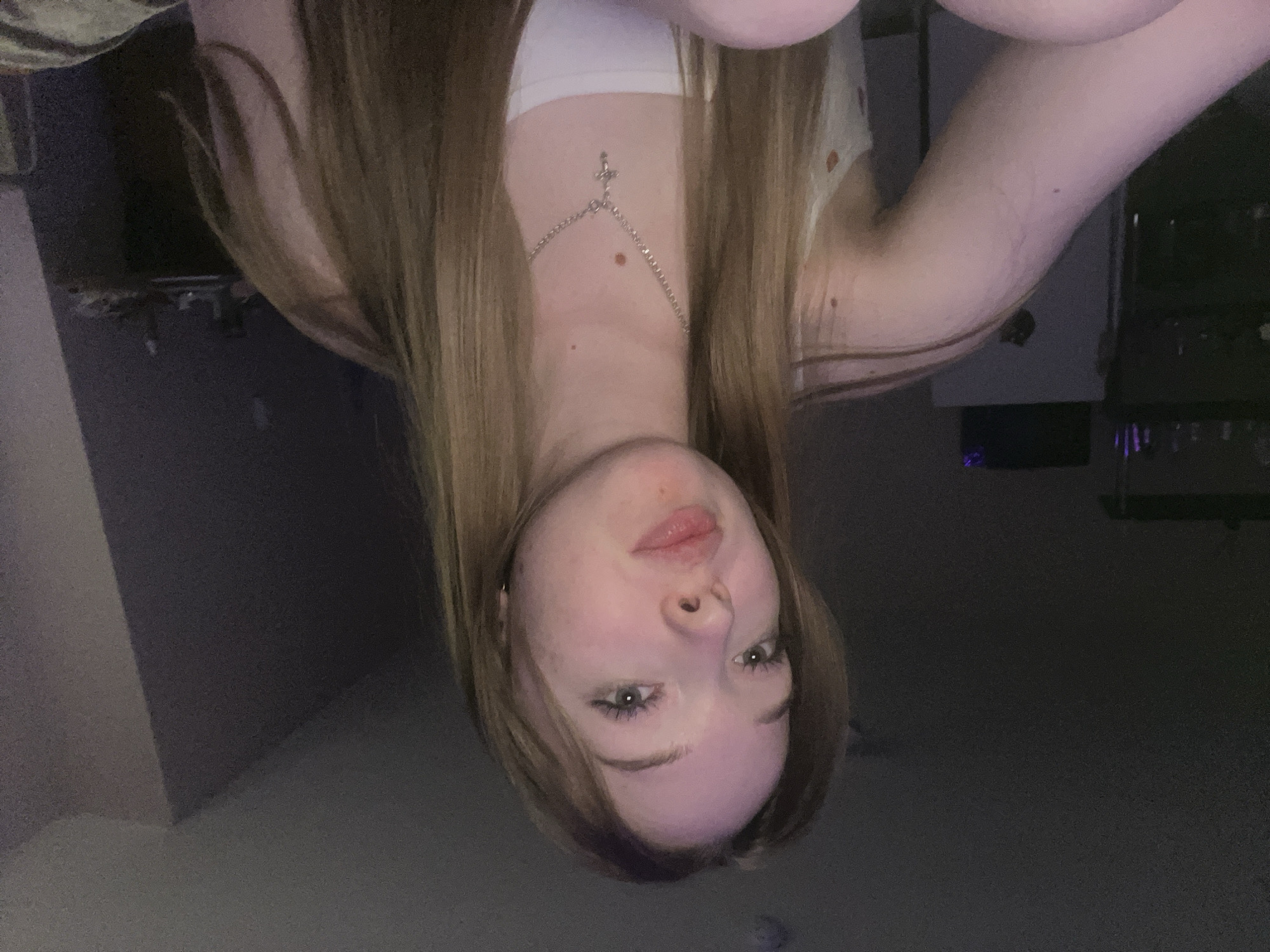 An informal portrait features a young person, likely female, with long, straight, light brown hair, looking directly into the camera. The person is positioned as if lying down, with their head tilted slightly upwards. Their expression is neutral to slightly pouting, with light-colored eyes. They are wearing a white or light-colored strapless top, and a silver chain necklace with a small cross pendant is prominent, resting on their upper chest. A couple of small moles are visible on their fair skin.
The background is very dark and out of focus, suggesting an indoor setting with low ambient light, possibly during the evening or night. Vague shapes in the background hint at furniture or room details, but nothing is clearly discernible. The primary light source illuminates the subject's face and upper body, creating a focused and somewhat intimate atmosphere. The image appears to be a casual selfie, possibly taken with a phone camera. There is no visible text. The scene is consistent with a personal space in Chaikovsky, Russia.