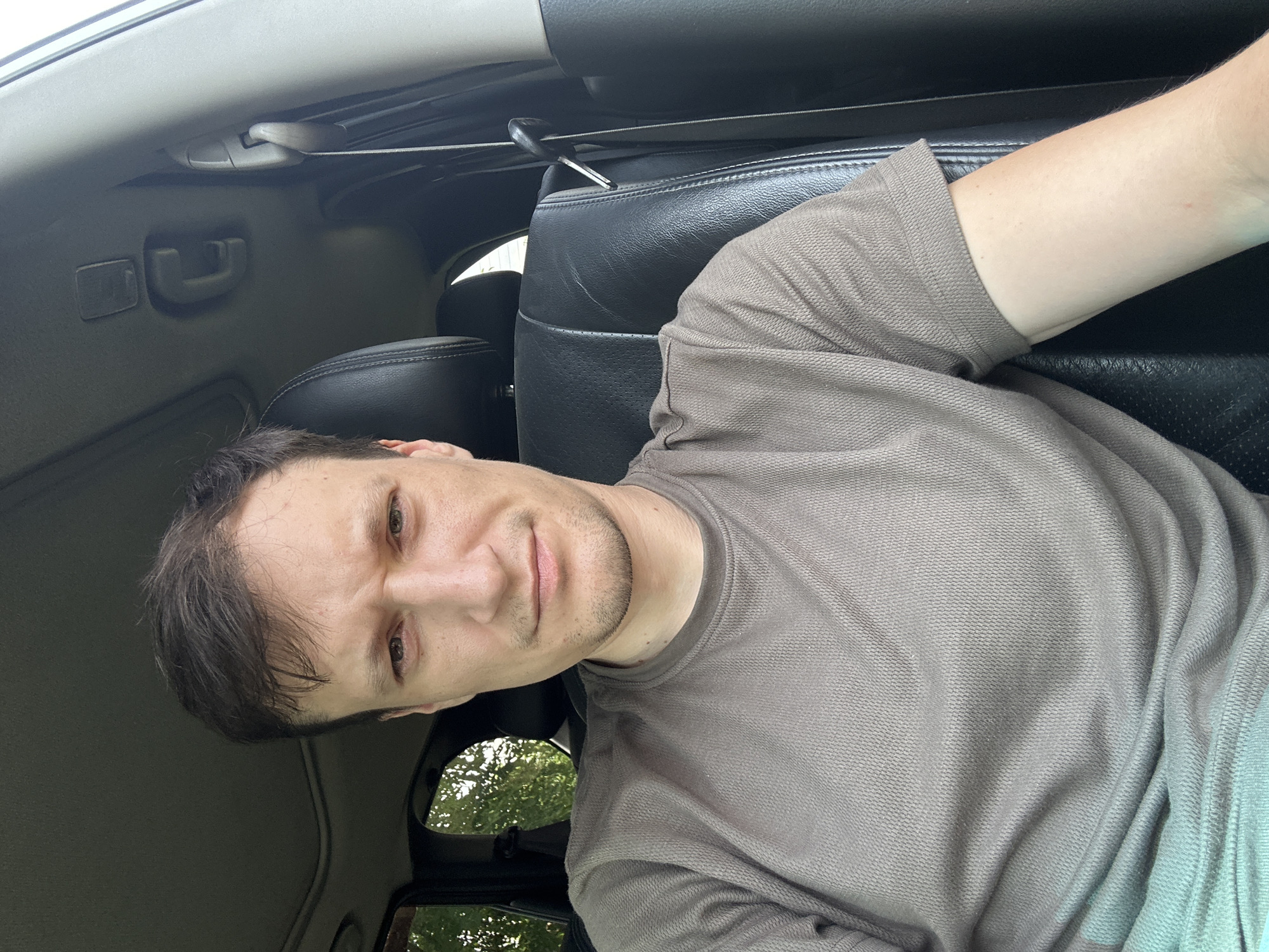 The image shows a man sitting in the driver's seat of a car. He is looking directly at the camera with a neutral expression. The man is wearing a short-sleeved, brown t-shirt. The interior of the car is visible, including black leather seats and a sunroof. Outside the car, trees are visible through the windows. The image appears to be taken during the daytime.