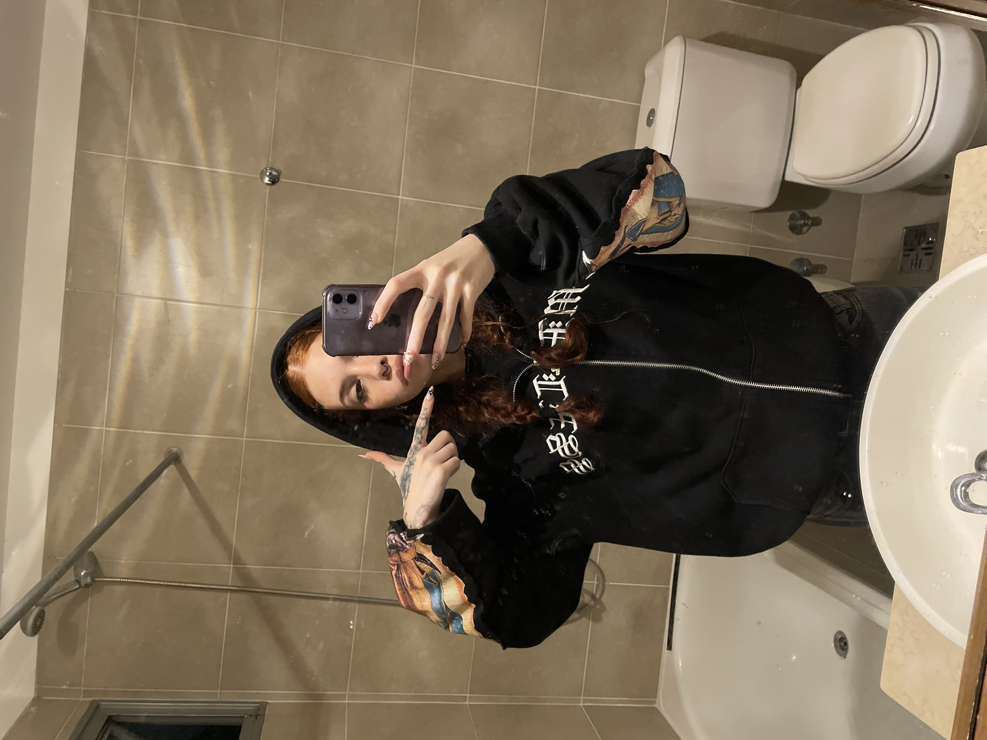 A selfie taken in a tiled bathroom, with the image rotated 90 degrees clockwise. A young woman with long, wavy reddish-brown hair, wearing a black zip-up hoodie, is reflected in the mirror. She holds a purple smartphone in her right hand, taking the picture, and points her left hand in a "finger gun" gesture towards the camera. Her nails are long and decorated, and tattoos are visible on her left arm. She has a nose piercing. The black hoodie features ornate white text, possibly in a Blackletter or Gothic font, on the chest area, and artistic prints resembling classical or renaissance paintings on the sleeves.
In the background, a light brown tiled wall forms the primary surface. A white toilet with its lid closed is positioned vertically on the right side of the mirror's reflection. Below the toilet, a white sink bowl and the edge of a counter are partially visible, along with a white bathtub or shower base. A metal shower rod structure extends horizontally across the left side of the frame, mounted on the tiled wall. Subtle light refractions are visible on the tiles. The scene suggests a casual, playful moment. The location context is Buenos Aires, Argentina.