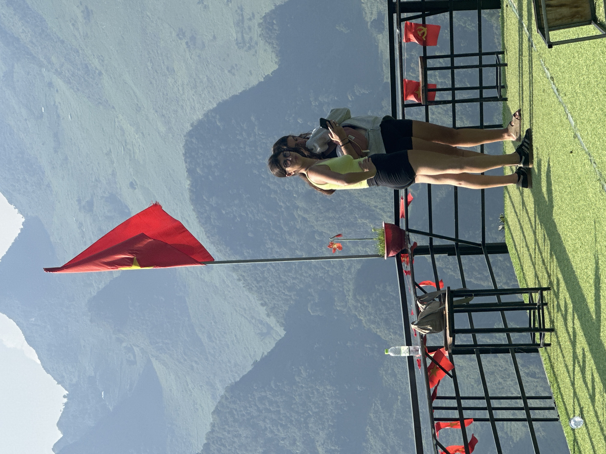 Two women stand on a grassy, elevated platform against a backdrop of green, mountainous terrain. The platform is constructed with black metal railings, adorned with small red flags, and also has several tables and chairs. One woman wears a bright yellow tank top and black shorts, while the other has a grey hoodie and black shorts. They are standing near a red flag, possibly Vietnamese, positioned prominently on a pole. The location appears to be in a mountainous area, possibly within or near Đường Thượng Commune, Vietnam, given the flags and the general environment.