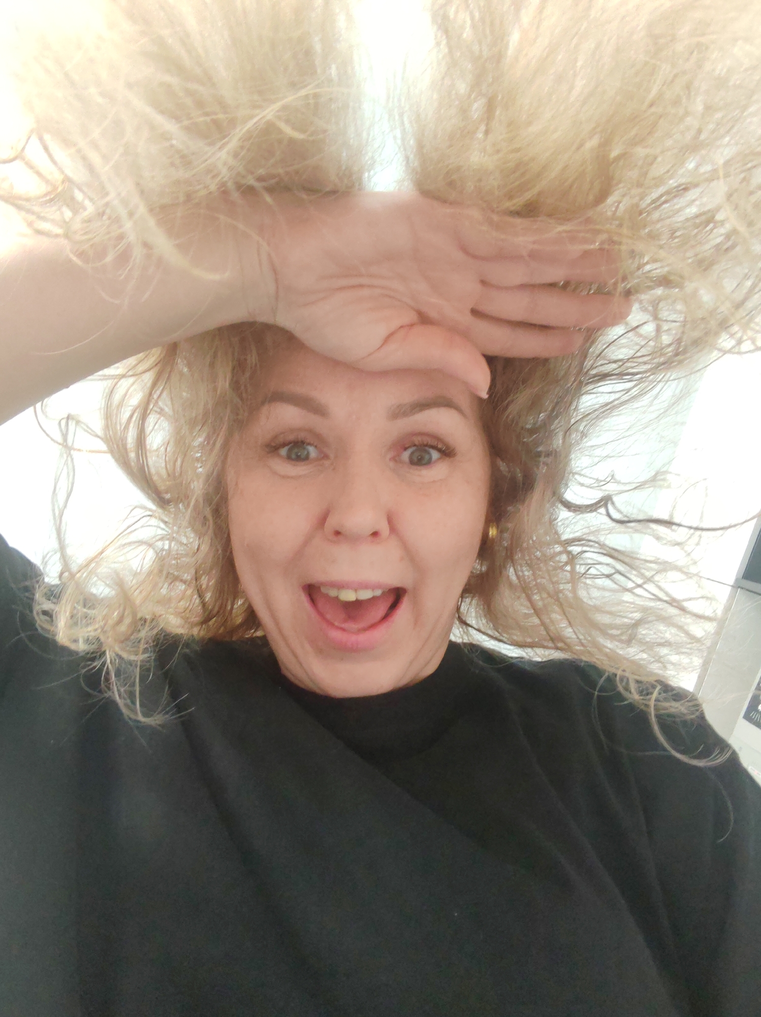 A woman with blonde, curly hair is pictured from a low angle, giving the impression that the viewer is looking up at her. She is wearing a black shirt. Her mouth is open in an expression of surprise or excitement, and she is holding her hand to her forehead as if shielding her eyes. The background is blurry and out of focus, but it appears to be an indoor setting. There is no information in the image to indicate that it was taken in the city of Chaikovsky, Russia.