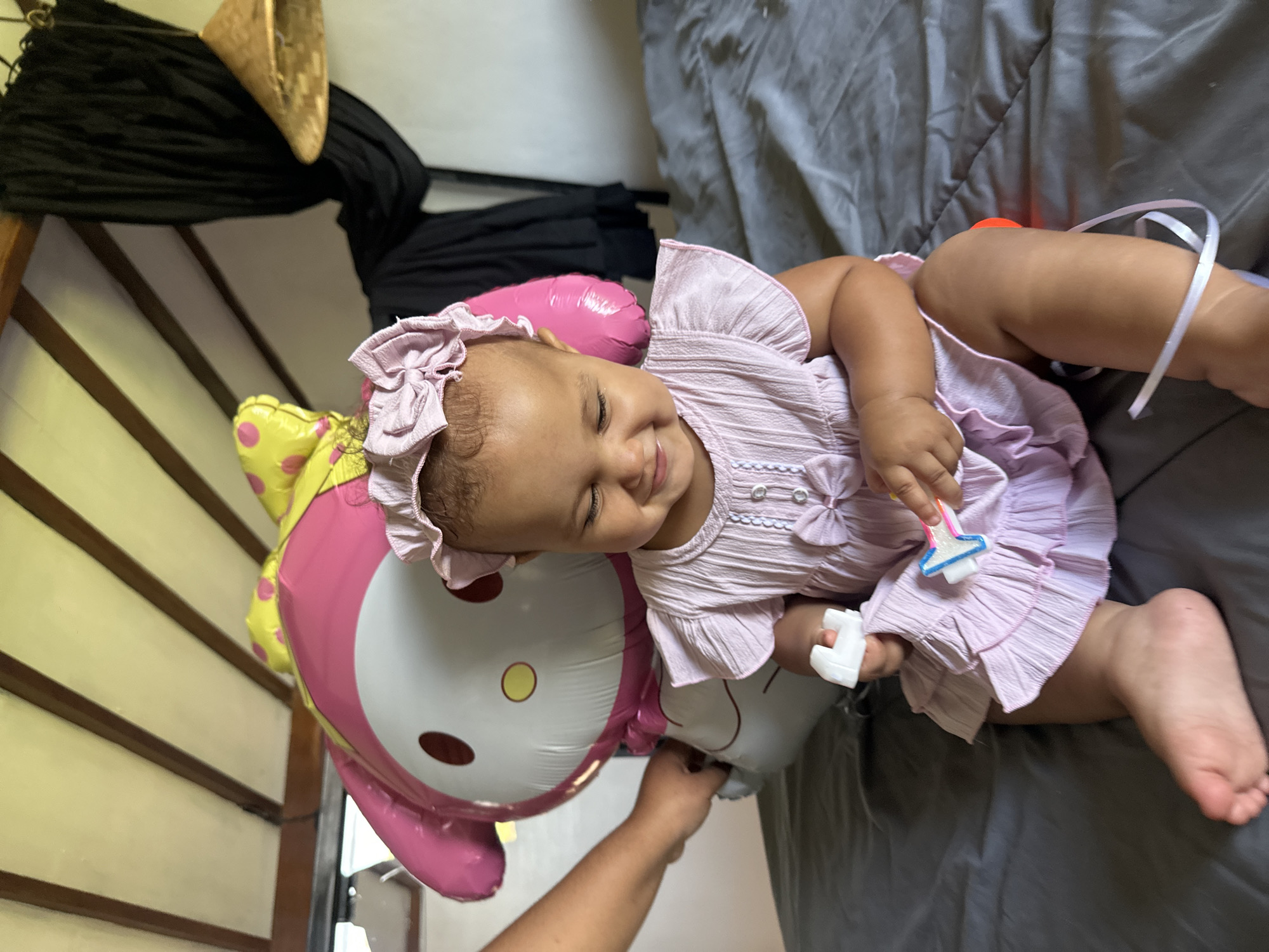 An infant girl, wearing a pink dress and matching headband, is seated on a bed with a grey comforter. She is looking down and smiling, holding two small toys in her hands. To her left, a large pink and yellow balloon in the shape of a character is partially visible. A hand is visible on the left side of the frame, holding onto the balloon string. The background shows a slanted wooden ceiling and dark drapery. The scene appears to be indoors, possibly in a bedroom.