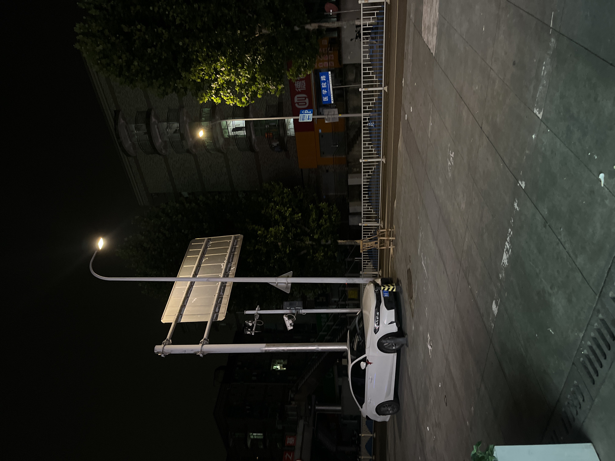 A white sedan is parked on a paved surface in front of a building at night. The car faces to the right, and its headlights are off. A street light illuminates the scene from the upper left. There are trees with green leaves on the right side of the image, and a building with multiple balconies is visible in the background. A white fence separates the paved area from the street. Signage in Chinese characters is present on the building and on a pole near the car. The ground is dark and appears to be made of concrete or asphalt, with some white markings and debris scattered on it. The sky is dark.