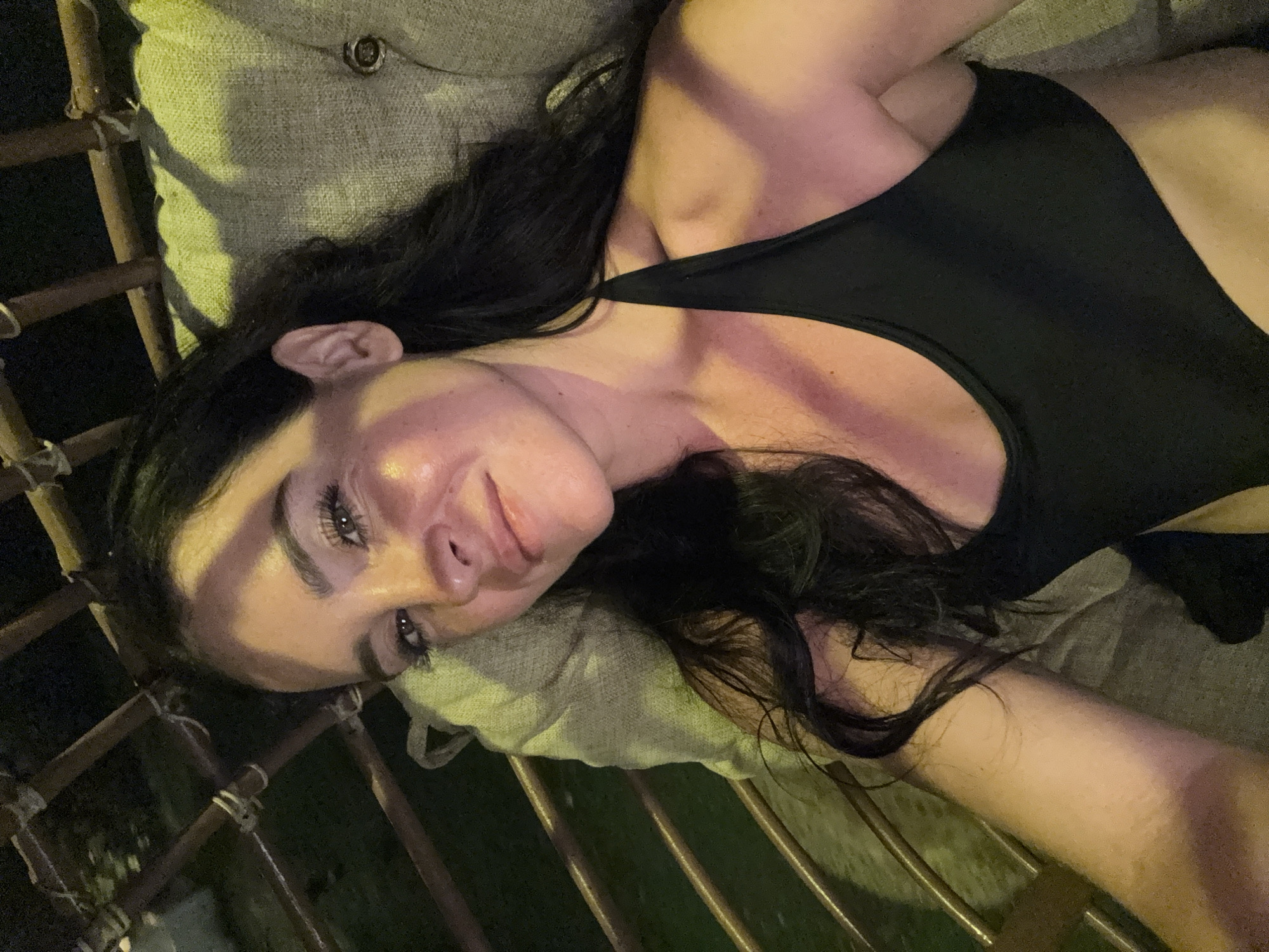 A young woman with long, dark hair and fair skin is lying down on a cushioned seat, possibly a hammock or swing chair. She is wearing a black halter top. Her face is turned towards the camera, and she is smiling slightly. The lighting is dim, with some artificial light illuminating her face and upper body. The background consists of dark bamboo-like poles and a green cushioned surface. The setting appears to be outdoors, likely at night. The location is identified as Karangasem, Indonesia.