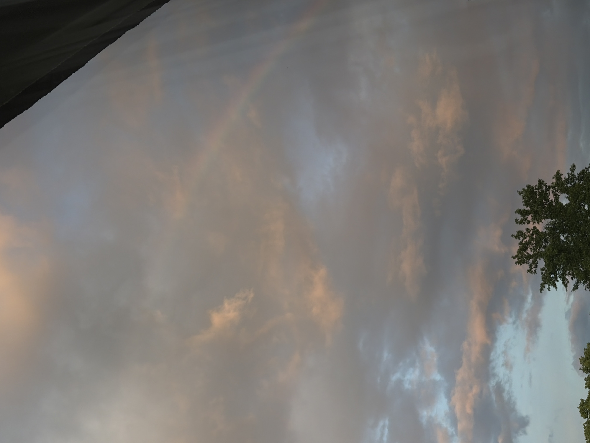 The image, oriented vertically, displays a cloudy sky with elements of a natural and potentially urban environment. The dominant feature is the expansive sky filled with diverse clouds, showcasing shades of grey, muted blue, and warm tones of pink and orange, suggesting either sunrise or sunset.
A faint, translucent rainbow arc is discernible in the upper-middle section of the image, subtly curving through the clouds. To the right, a cluster of vibrant green tree foliage is visible, indicating the presence of trees in the foreground or midground. A dark, diagonal structure, possibly a roofline or part of a building, occupies the top-left corner, suggesting a man-made element in the scene.
The clouds range from dense, darker formations to lighter, more ethereal wisps. No human activity or specific events are depicted, and there is no visible text. The atmosphere is serene and natural, highlighted by the interplay of light and color in the sky.