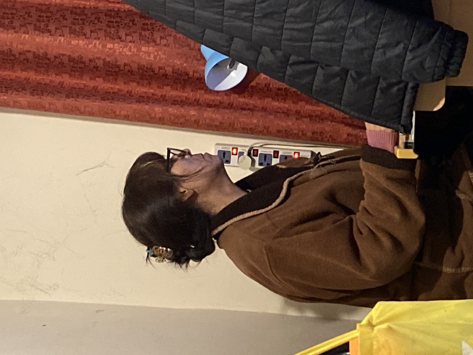 An individual is depicted indoors, in what is identified as a location within La Paz, Bolivia. The person, appearing to be an adult female, is shown in left profile, seated or positioned behind a surface. She has dark hair styled in a low bun, secured with a decorative clip, and wears black-framed eyeglasses. Her attire includes a thick, brown fleece-like jacket with a darker collar and trim. A light pink cuff is visible on her right wrist.
The immediate background consists of an off-white wall with visible hairline cracks. Mounted vertically on this wall is an electrical power strip with multiple outlets, several of which display illuminated indicators. To the right of the power strip, a patterned red curtain is visible. In the right mid-ground, a light blue and white lampshade is partially visible, obscured by a dark, quilted jacket hanging or resting on an unseen object. In the lower left foreground, a portion of a yellow bag or container is visible. The individual's hands are positioned near a light-colored surface, possibly a desk or a keyboard, with yellow-colored keys or controls partially discernible. The overall scene suggests an indoor, domestic, or workspace environment with artificial lighting.
