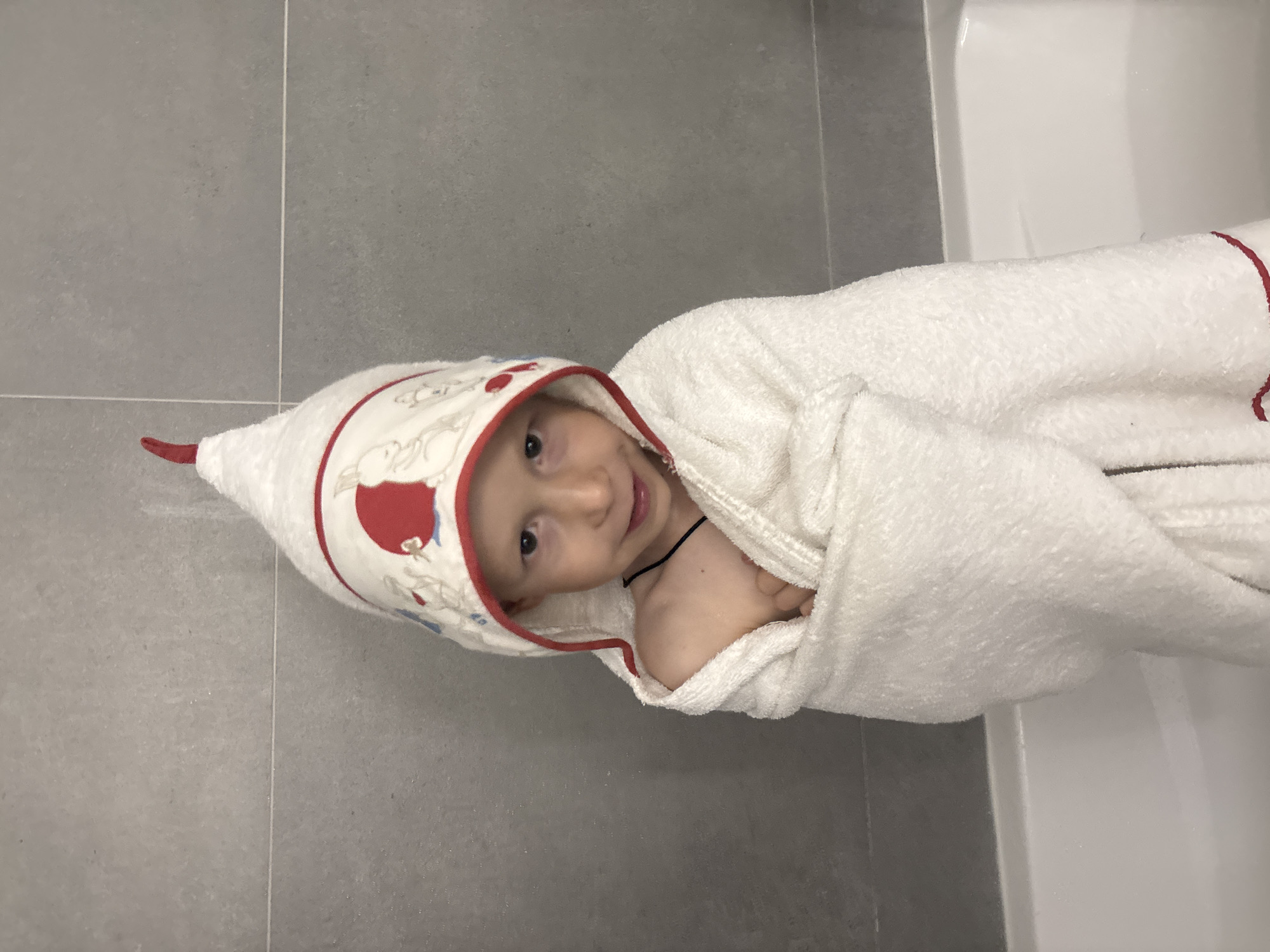 A young child, wrapped in a hooded white towel with red trim and holiday-themed decorations, lies on a gray tiled floor adjacent to a white bathtub. The location is consistent with a domestic bathroom in Chaikovsky, Russia. The child appears to be post-bath. The towel is wrapped around the child's body and head.