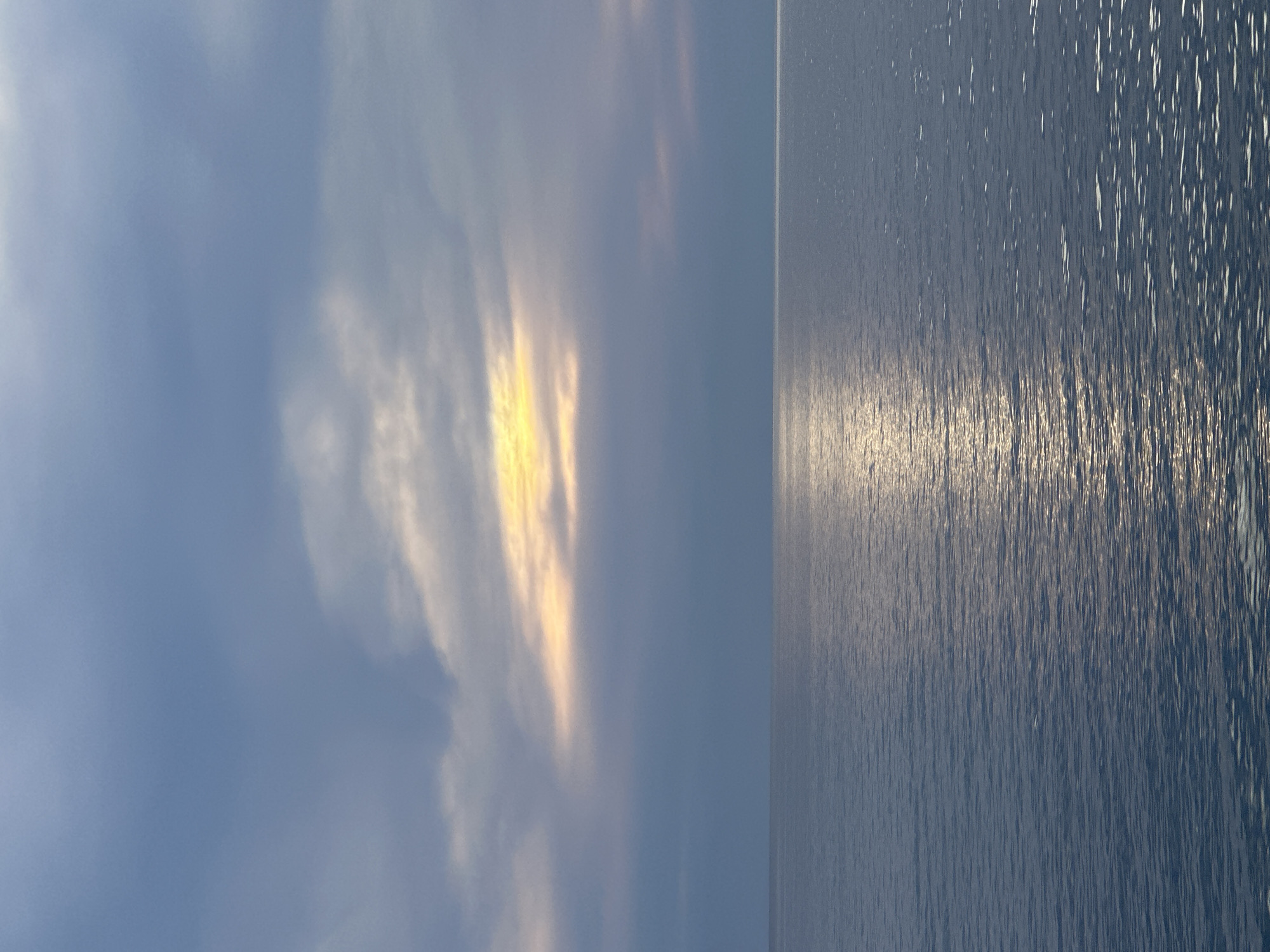 The image shows a split view of the sky and the sea, likely taken in Bar, Montenegro. The sky is predominantly blue with scattered white and yellow clouds, indicating either sunrise or sunset. The sea is calm, with the sun's reflection creating a shimmering effect on the water's surface. There is no visible human presence or man-made objects, creating a naturalistic landscape scene.