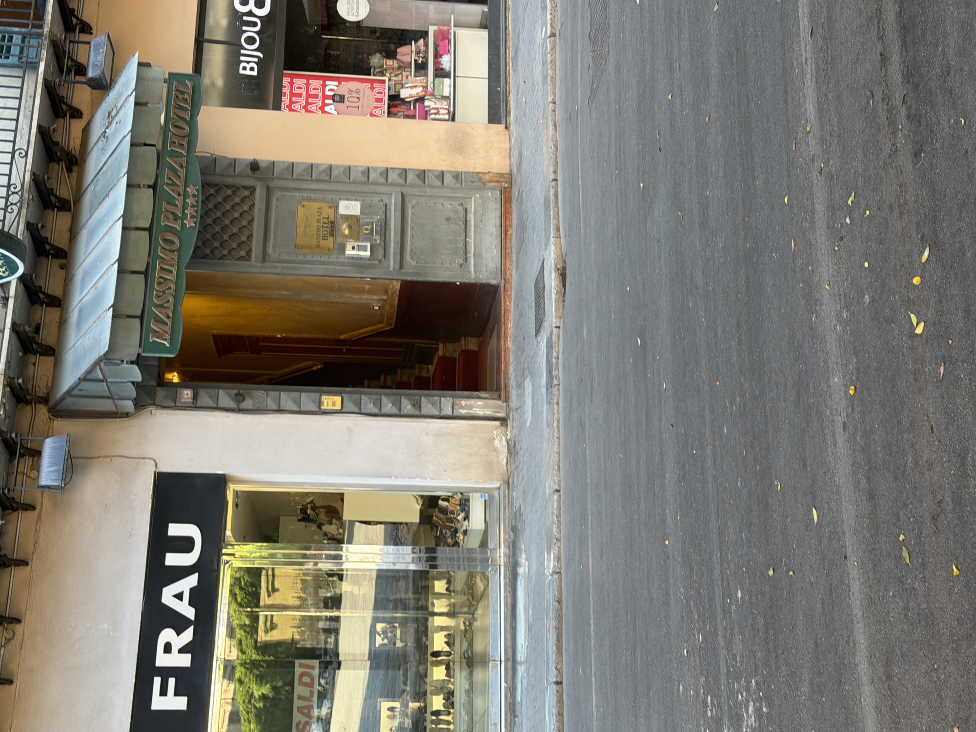 This is a street-level exterior view of shops and a hotel entrance in Palermo, Italy. On the left, a store with a large black sign reading "FRAU" is visible. The store features a glass storefront displaying shoes and other merchandise. To the right of "FRAU" is the entrance to the "MASSIMO PLAZA HOTEL," marked by a green awning and a sign with four stars. The hotel door is grey and has a metal plaque. Further to the right, a shop window displays a pink jacket and bags, with signs for "ALDI" and "BIJOU." The street in the foreground is paved and has fallen leaves scattered across it. The architecture suggests a European city setting.