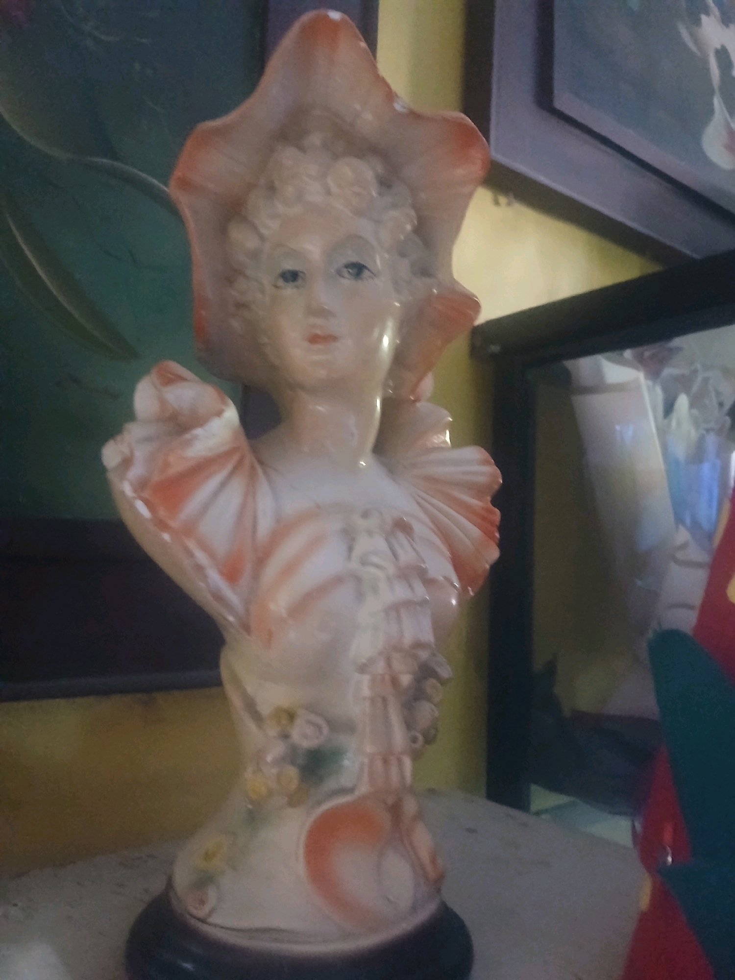 A ceramic figurine of a woman wearing a large hat and elaborate dress is displayed indoors. The figurine is light-colored with pink and white accents, featuring delicate facial features and floral embellishments on the dress. The background shows a dark green object to the left and framed artwork or photographs to the right, with a yellow wall behind. The scene is dimly lit, suggesting an indoor setting likely within a home or shop. The figurine is positioned on a surface that appears to be a shelf or table, with a dark circular base. No people are visible in the image, and no specific location in Banyuwangi, Indonesia, can be determined from the provided visual information.
