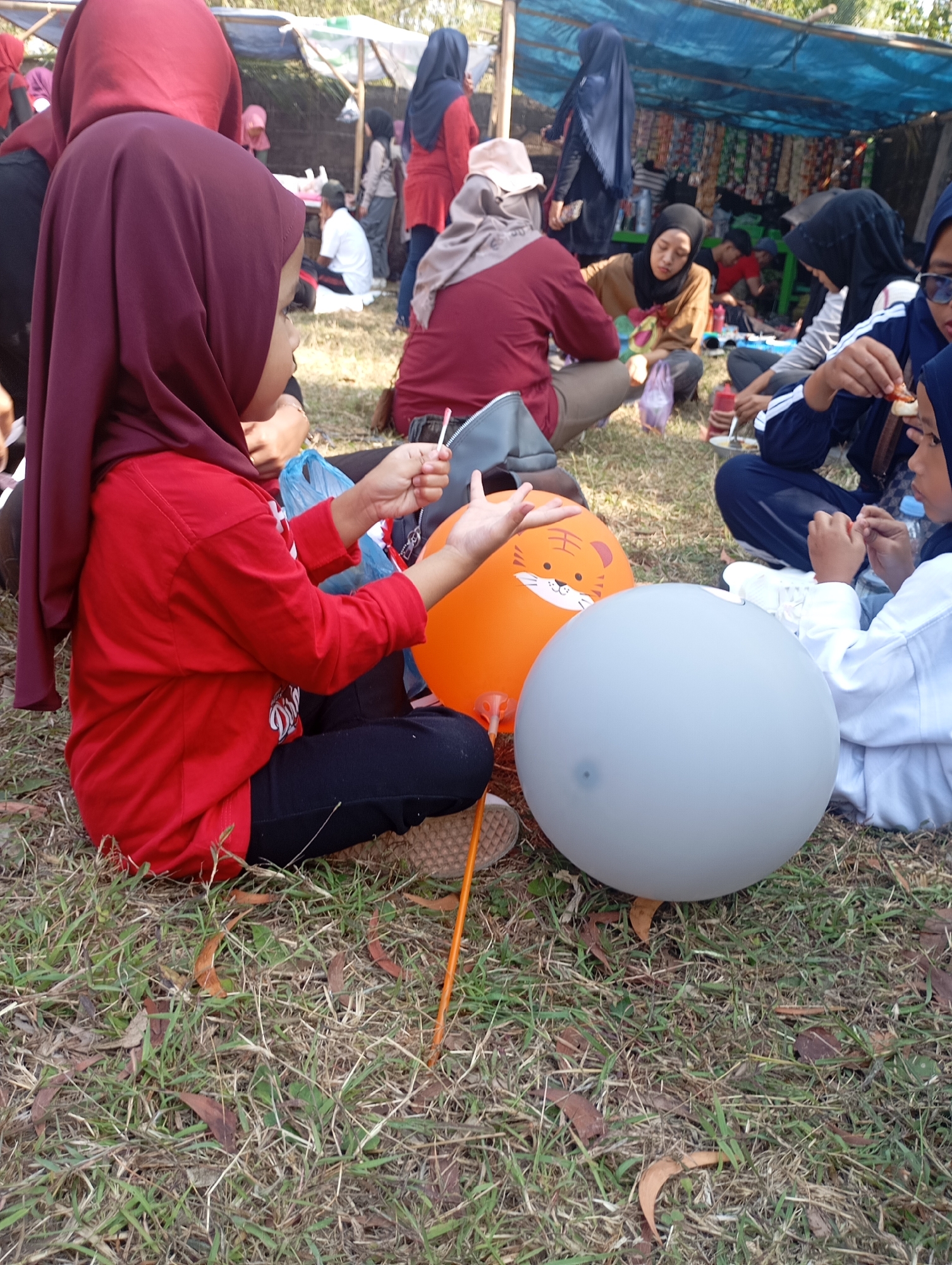 In Ketepung, Indonesia, a group of individuals, predominantly young females wearing hijabs and modest attire, are gathered outdoors on a grassy area. In the foreground, a young girl in a red shirt and black pants is seated, holding a lollipop and reaching towards balloons. Two balloons are visible: an orange one with a tiger face print and a large, light gray one. Other individuals are scattered around, some seated and engaged in eating or conversation, while others are standing near a makeshift stall covered by a blue tarp. The environment appears to be a casual gathering, possibly a community event or picnic, with fallen leaves and dry grass on the ground.