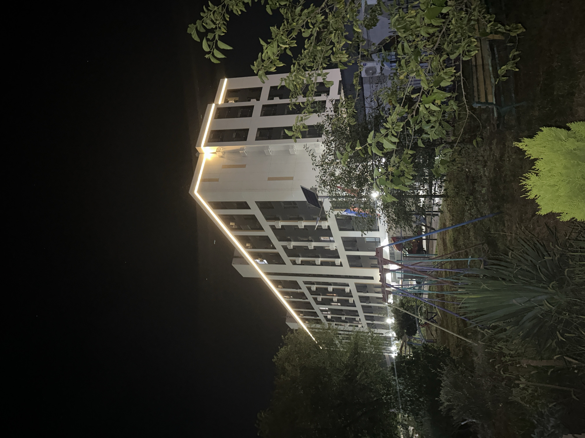 The image shows a modern, multi-story apartment building at night, illuminated by a string of warm white lights along its roofline. The building has a white facade and dark windows arranged in a grid pattern. In the foreground, there is a children's playground with swings and a metal fence. Dense foliage from trees and bushes obscures parts of the scene. The ground is covered in grass and dirt. The overall atmosphere is quiet and still, suggesting a residential area. The location is identified as Nevinnomyssk, Russia.