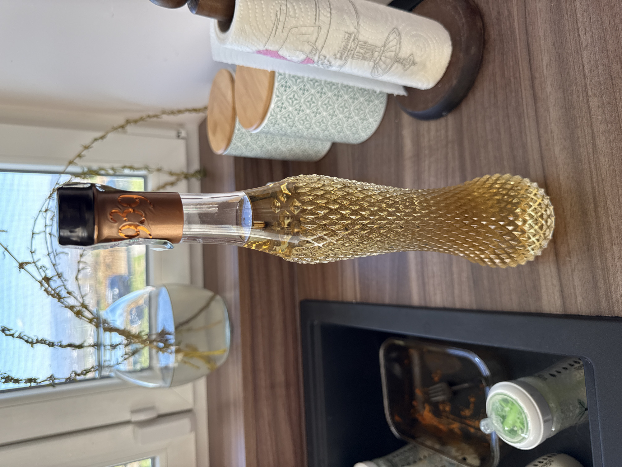 On a wooden countertop, an elongated glass bottle with a textured golden-yellow body and a black cap lies horizontally. The bottle is positioned next to a window where a glass vase with thorny branches is visible. Above the bottle, there's a paper towel holder and a decorative storage container. Below the bottle, a black sink contains a metal tray and a bottle with a green cap. The scene is well-lit, suggesting an indoor environment. The location is Sofia, Bulgaria.