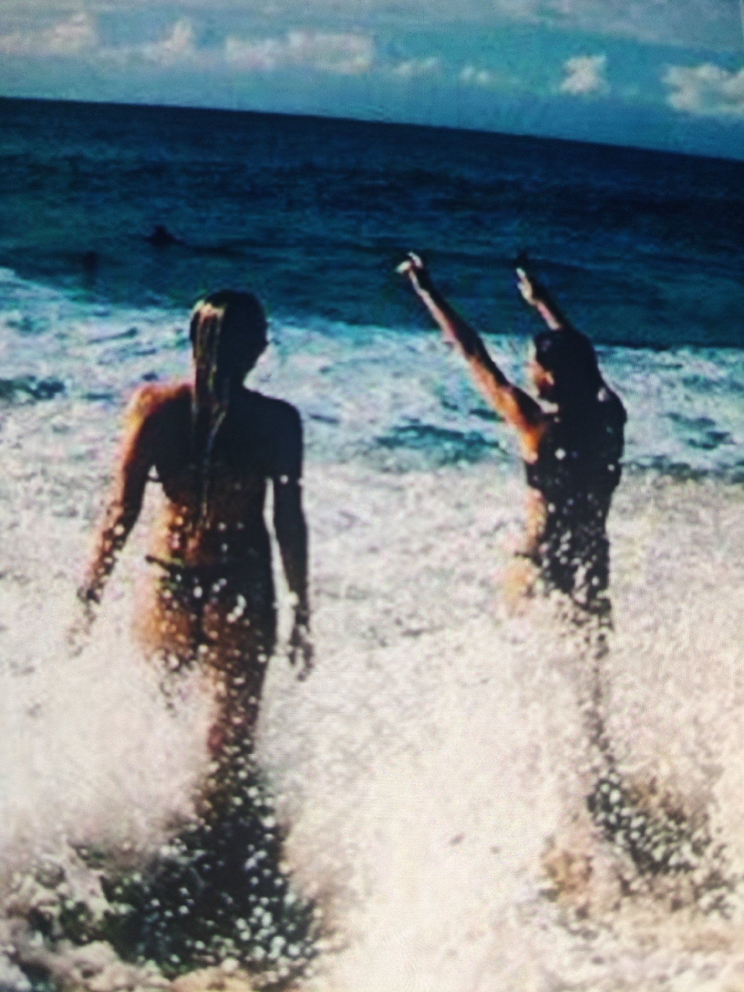 The image depicts two individuals in the ocean. One person is facing away from the camera, wading through the water. The second person is facing the ocean with their arms raised in the air. The water is choppy and appears to be breaking into waves around them. The sky above is partly cloudy. There is no indication of the city of Krasnaya Polyana, Russia, in this image.