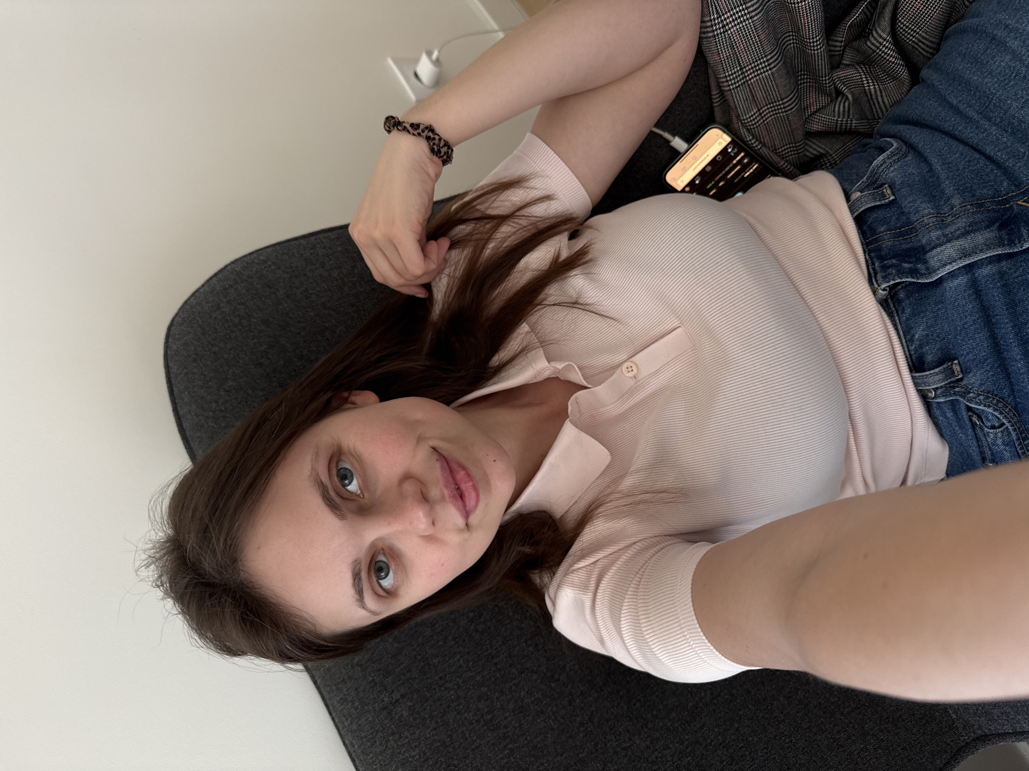 A young woman is lying on a dark gray couch, presumably indoors. She has fair skin, brown hair, and blue eyes. She is wearing a light pink, short-sleeved, collared shirt and blue jeans. A brown scrunchie is on her wrist. A smartphone is plugged in and lying beside her on the couch. Part of a plaid jacket is visible next to her jeans. The wall behind her is white, and an electrical outlet is visible. There are no obvious indicators of the location being Belgrade, Serbia.