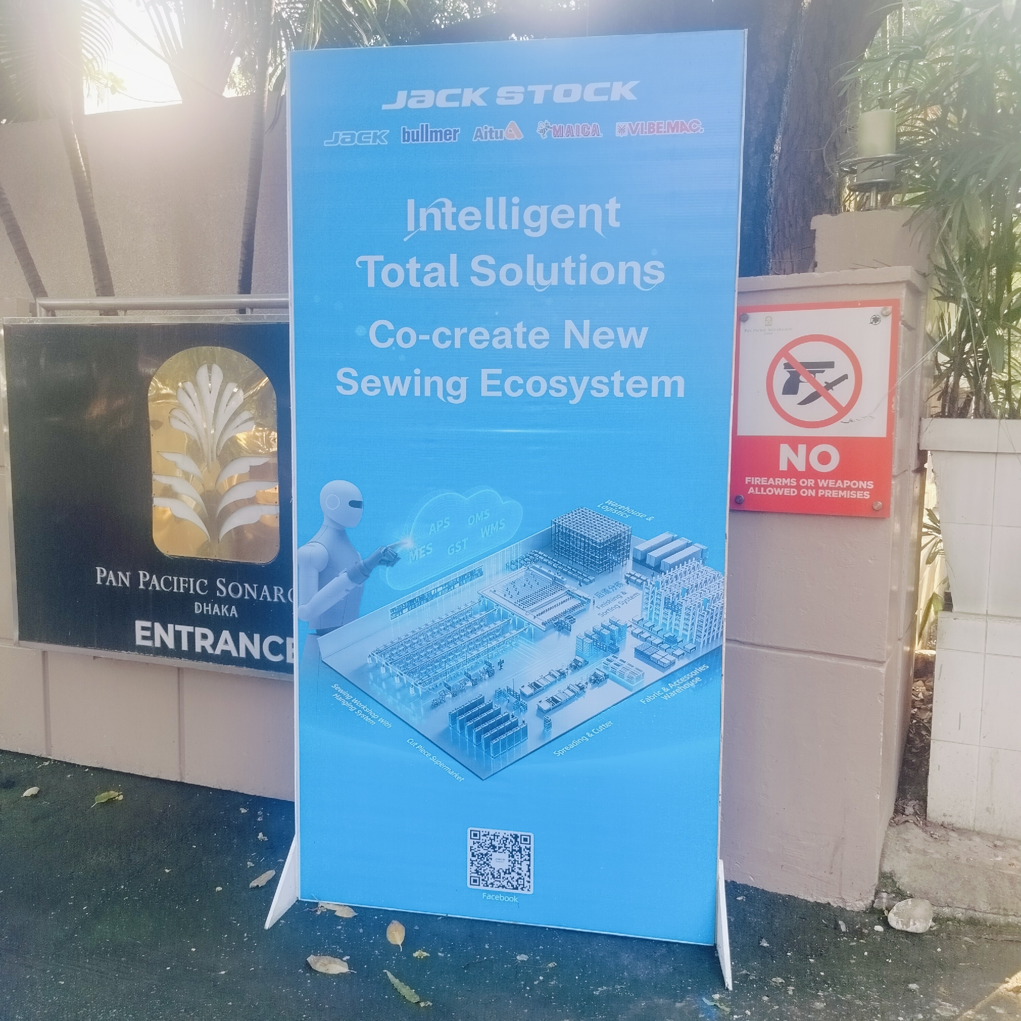 The image shows a stand-up advertisement for JACK STOCK sewing solutions. The sign is blue and features the company's name, slogans, a robotic arm reaching toward a diagram of a sewing ecosystem, and a QR code. The sign is positioned next to the entrance sign for the Pan Pacific Sonargaon hotel in Dhaka. A "No Firearms or Weapons Allowed on Premises" sign is attached to the adjacent wall. The ground appears to be a dark, possibly wet surface.