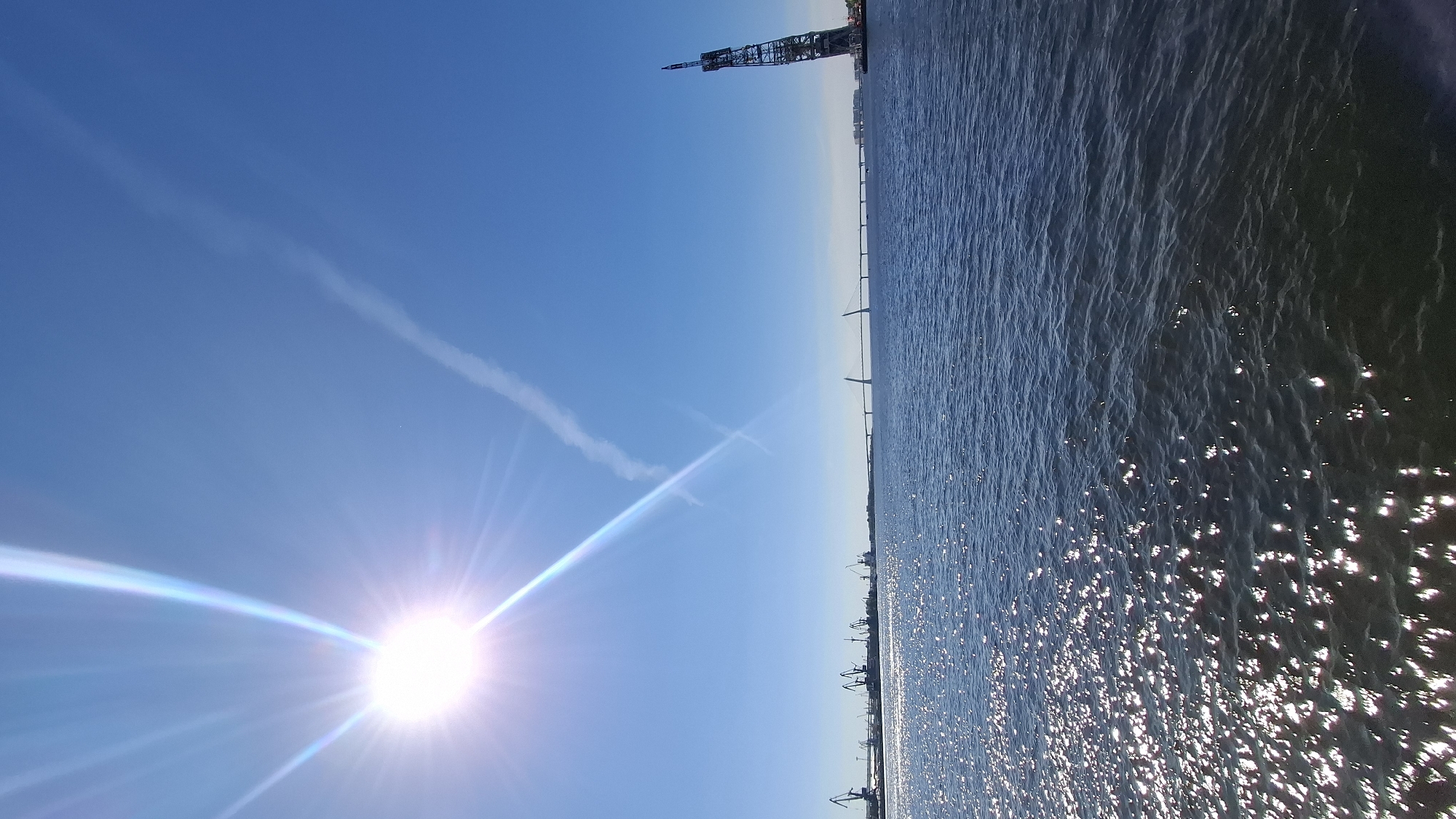 The image depicts a wide body of water under a clear blue sky with the sun prominently shining in the upper left corner, casting bright rays. In the distance, a long bridge spans across the water, with two distinct towers visible on the right side. To the left, a line of cranes indicates a port or industrial area. The water's surface is choppy, with sunlight glinting off the waves, creating a sparkling effect. A faint contrail is visible in the sky. The scene is characteristic of a waterfront view in Saint Petersburg, Russia, likely the Neva River or a bay area, given the presence of port infrastructure and a large bridge. There are no people visible in the frame.