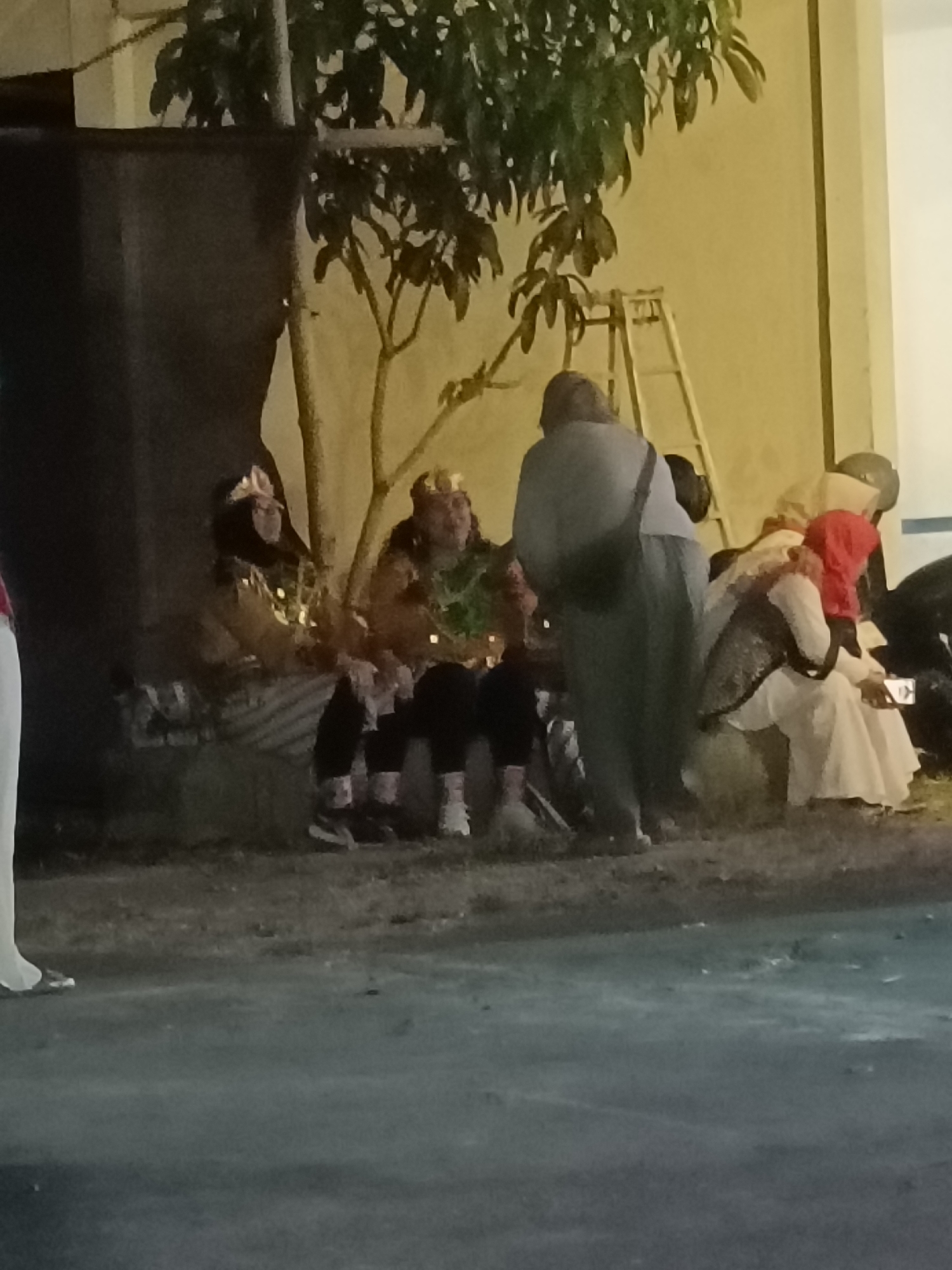 The image depicts a group of people gathered outdoors, possibly at night or in low-light conditions, in what appears to be a public space in Kediri City, Indonesia. In the foreground, the ground is a mix of grass and asphalt. A dark, rectangular object, possibly a barrier or a stage, is visible on the left. Behind this, a tree with dark green leaves is prominent, with a ladder leaning against a light-colored wall in the background.
Several individuals are seated on what seems to be a low, dark bench or ledge. On the far left, a person in a white outfit is partially visible. Next to them, a person wearing a dark outfit and a head covering is seated, adorned with what look like small, decorative lights. To their right, another person in a similar dark outfit is seated, wearing a crown-like accessory and holding an object that may be a prop or a musical instrument. Further to the right, a person in a dark gray outfit is standing, facing away from the camera, and appears to be wearing a bag over their shoulder.
To the right of the standing person, a group of individuals are seated. One person wears a light-colored headscarf and a patterned garment. Another, in the foreground of this group, wears a red headscarf and a white dress, holding a small rectangular object, possibly a phone. The overall impression is of a community gathering or a performance taking place outdoors in Kediri City.