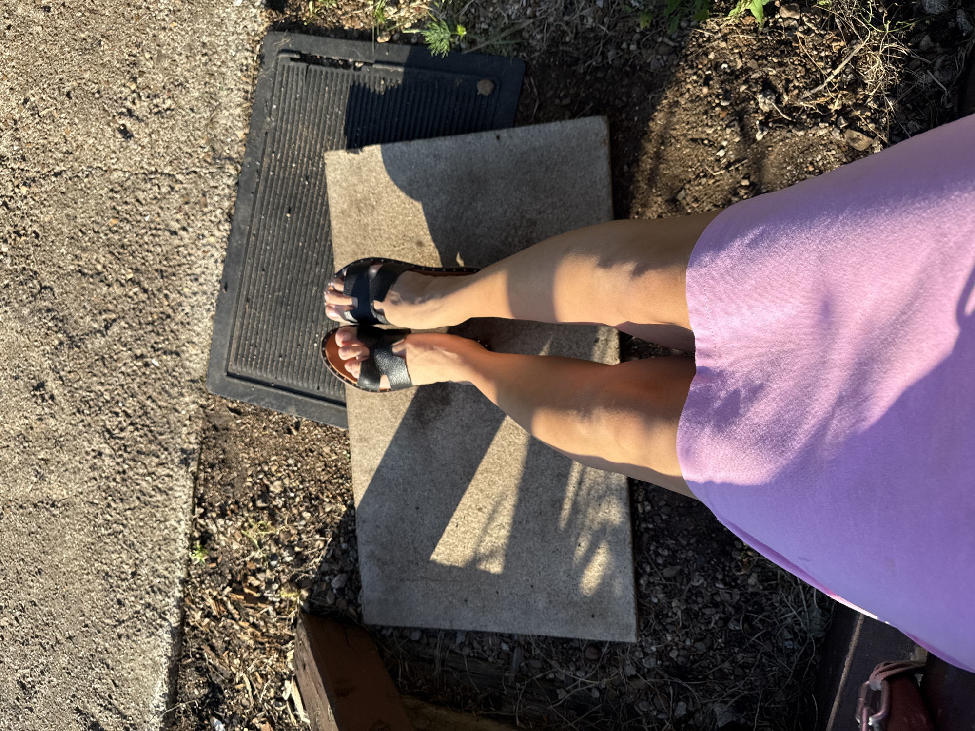 A first-person perspective shot captures a person's legs and feet from above, seated on a wooden bench. The person is wearing black sandals and a purple skirt. The ground is a mix of dirt, gravel, and wood chips. In the background, a concrete slab and a black rubber mat are visible. The scene is illuminated by bright sunlight, casting distinct shadows. The location appears to be an outdoor setting, potentially a backyard or a park area within Verkhneyarkeyevo, Russia.