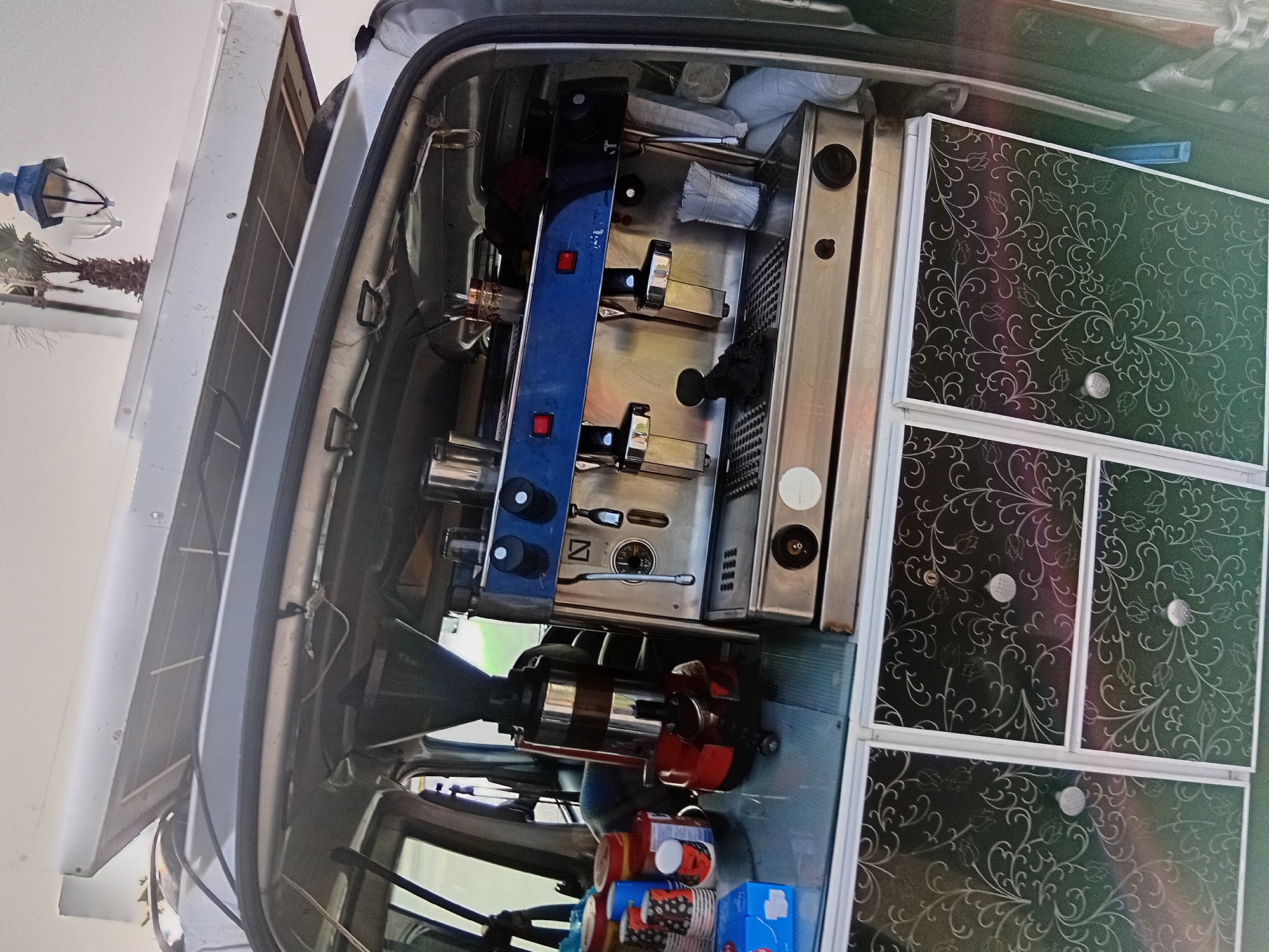 This media file captures a custom-built mobile coffee setup inside a vehicle, likely a van or food truck, designed for operating an outdoor coffee business. The image is taken from a slightly elevated angle, looking down into the vehicle's interior.
The central focus is a professional-grade espresso machine, prominently featured with a blue front panel, stainless steel accents, and multiple group heads. It has red and black control buttons, a pressure gauge, and steam wands. To the left of the espresso machine is a substantial coffee grinder with a red base and a black hopper, positioned on a lower counter.
On the right side of the vehicle's interior, there are several dark-colored cabinets or drawers with intricate white or silver floral/swirl patterns on their doors and small silver knobs. Below the main counter, various supplies are visible, including stacks of colorful paper cups with patterns like red polka dots, and blue boxes or containers, suggesting provisions for serving beverages.
Outside the vehicle, a large solar panel is mounted on the roof, indicating an off-grid power solution for the coffee equipment. In the background, beyond the vehicle, parts of a white lamppost and the top of a palm tree are visible against a bright, clear sky, suggesting daytime and good weather conditions. No people are visible, but the setup implies the imminent or ongoing activity of a mobile coffee vendor. The scene is likely set in Martil, Morocco, as per the provided context. No discernible text is visible on any items.