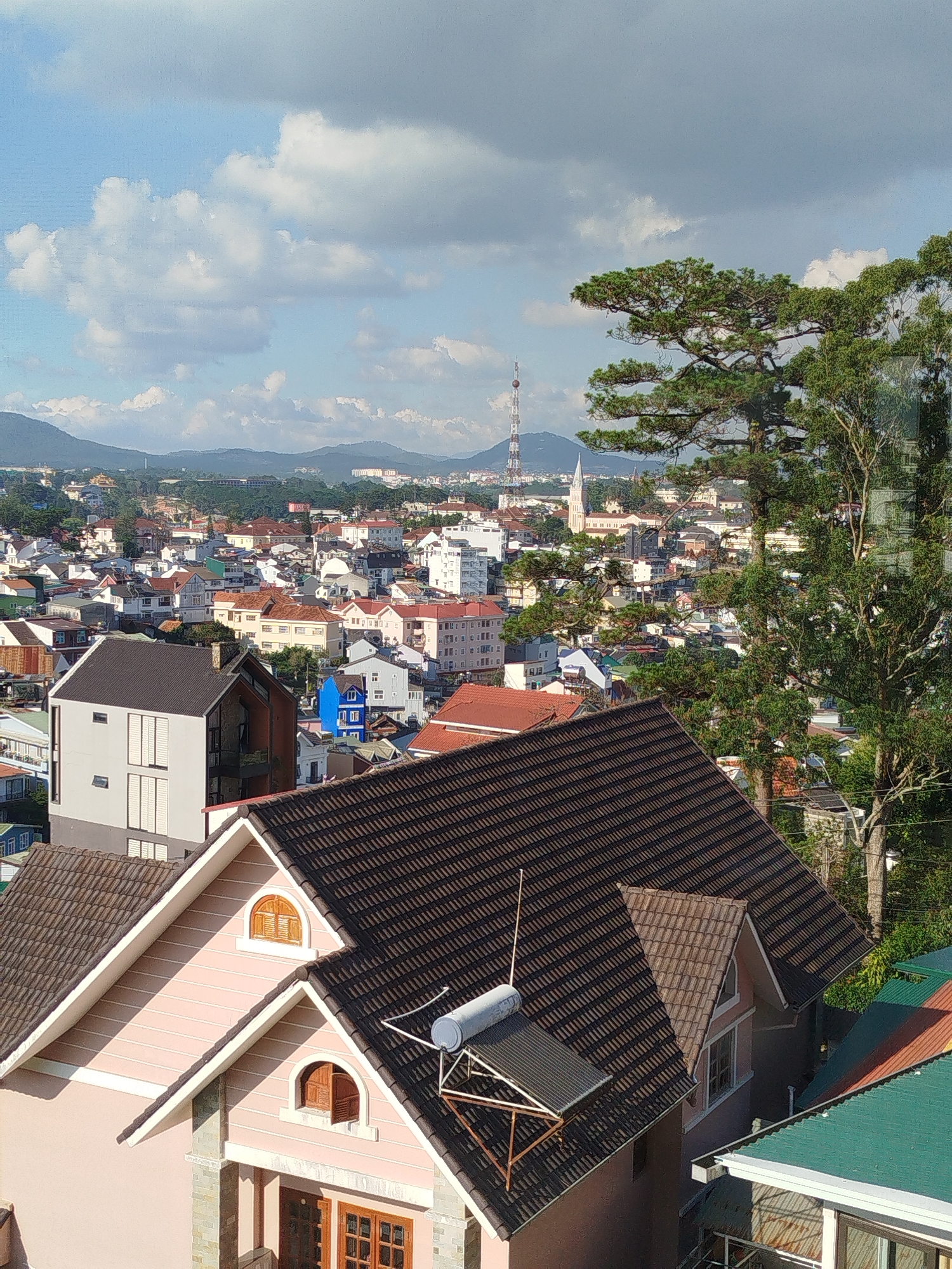 The image presents an elevated view of Da Lat, Vietnam, characterized by a dense urban landscape and rolling hills in the background. The foreground is dominated by several buildings with distinctive tiled roofs, predominantly brown and green, indicating a residential area. A pink building with a brown tiled roof features arched windows and a solar water heater on its roof. In the midground, a mix of building styles and colors is visible, including white, grey, and blue structures. A radio tower and a church steeple are prominent landmarks against the skyline. The background reveals a series of mountains under a sky dotted with puffy white clouds. Trees are visible on the right side of the frame, adding a touch of greenery to the scene. No people are visible in the image.