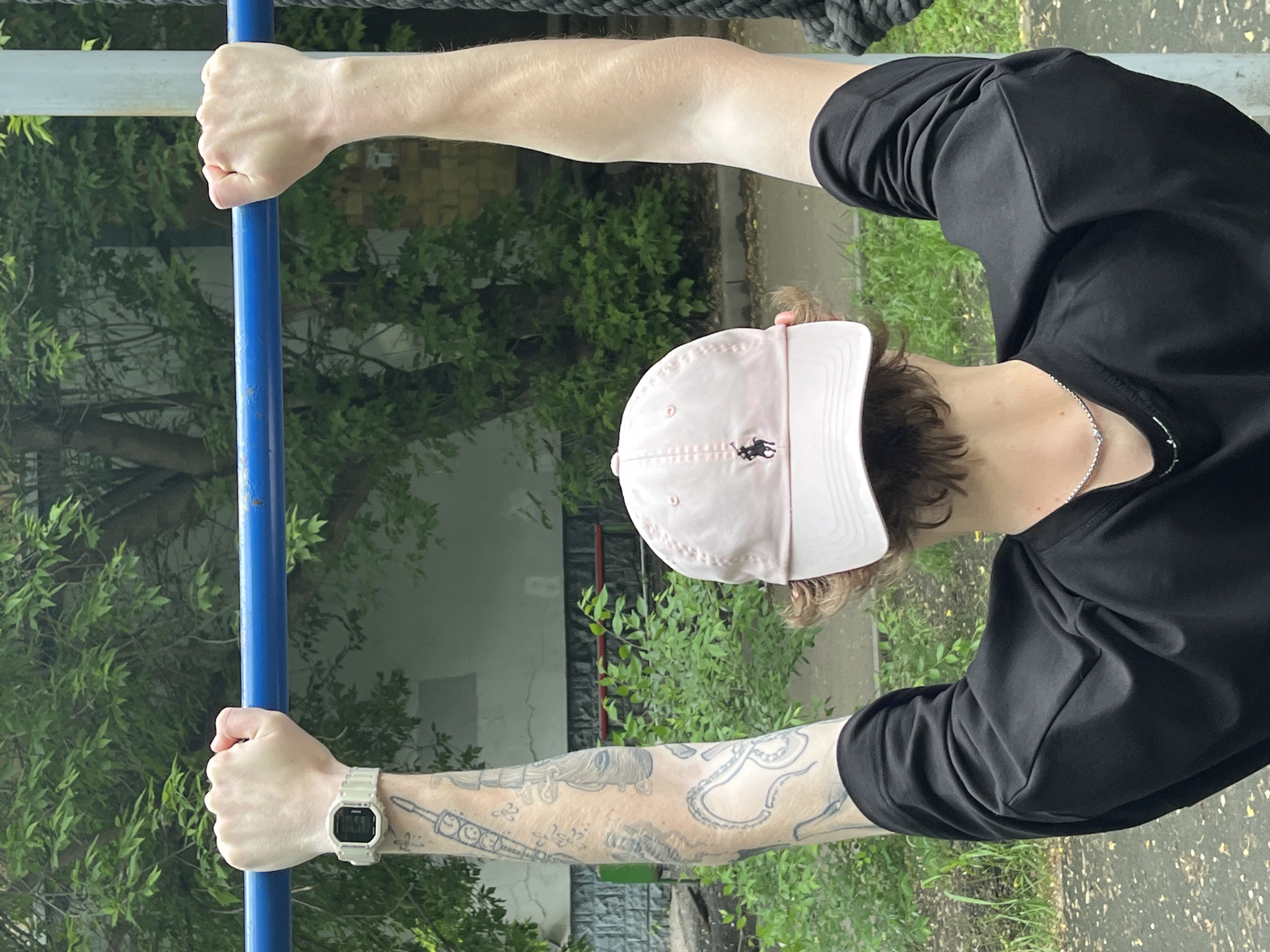 A person is seen from the back, performing a pull-up on a blue horizontal bar in an outdoor urban setting. The person is wearing a black long-sleeved shirt, a light pink baseball cap with a black logo, and a silver necklace. A watch is visible on their left wrist, and tattoos are present on their left arm. The background consists of green foliage, trees, and a building. The ground appears to be asphalt or concrete. The scene is likely located in Krasnoyarsk, Russia, as indicated by the specific instructions.