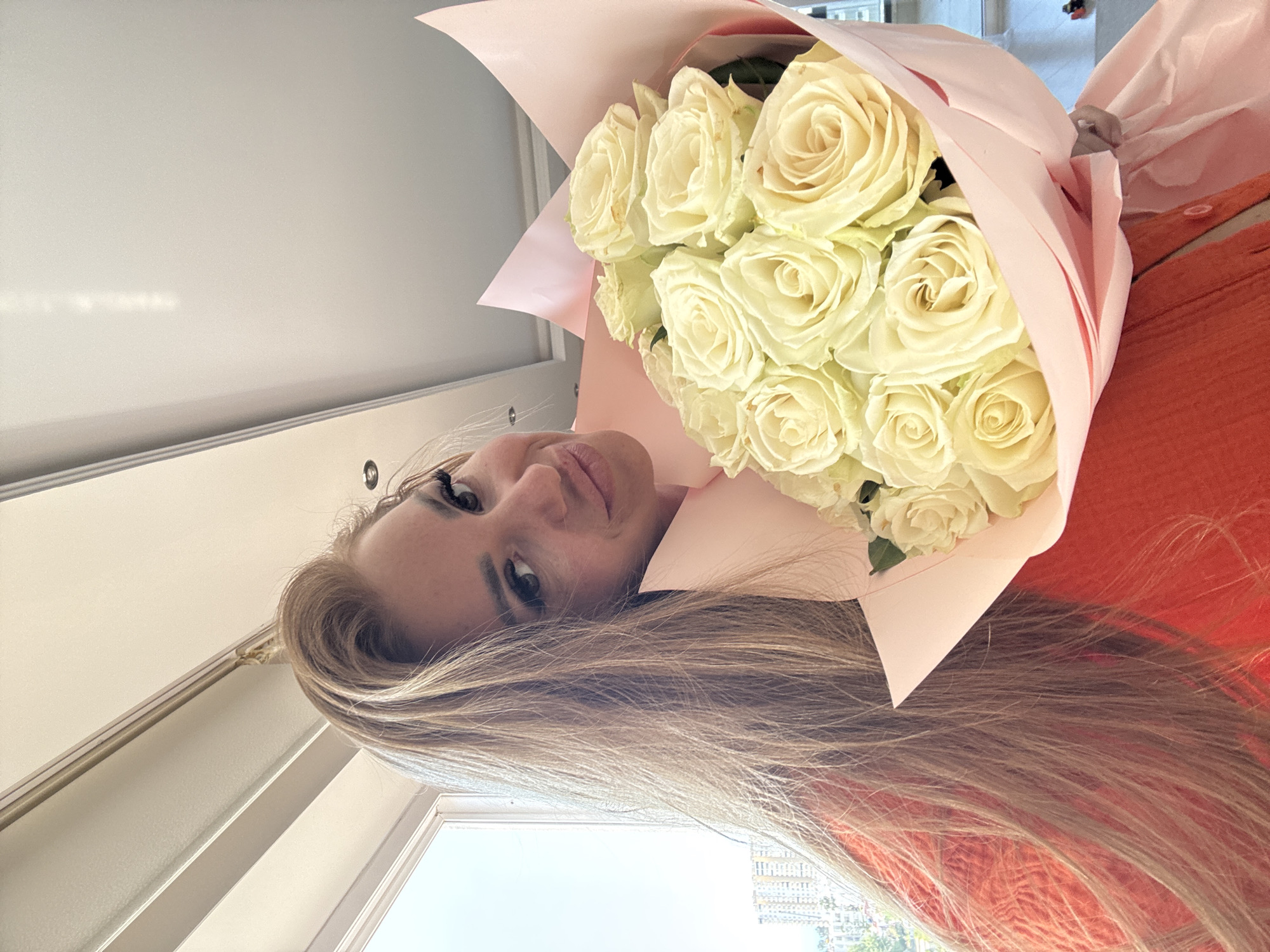 A close-up, upward-angled shot captures a woman holding a large bouquet of white roses. The woman, possibly in her 20s or 30s, has long, light brown hair that cascades towards the bottom of the frame. Her face is partially visible, featuring dark eyes looking towards the camera and slightly parted lips, conveying a neutral to pleasantly content expression. She is wearing an orange-red garment, with a hint of a light pink top or scarf around her neck area.
The bouquet, wrapped in delicate light pink paper, consists of numerous creamy white roses, their petals fresh and perfectly formed. The flowers are held prominently in the foreground. In the upper part of the image, a plain white ceiling is visible. Towards the lower-left, a window offers a blurred view of the outside, revealing faint outlines of buildings and some greenery, suggesting a bright daytime setting in an urban or suburban location like Erdemli, Turkey. There is no visible text in the image.