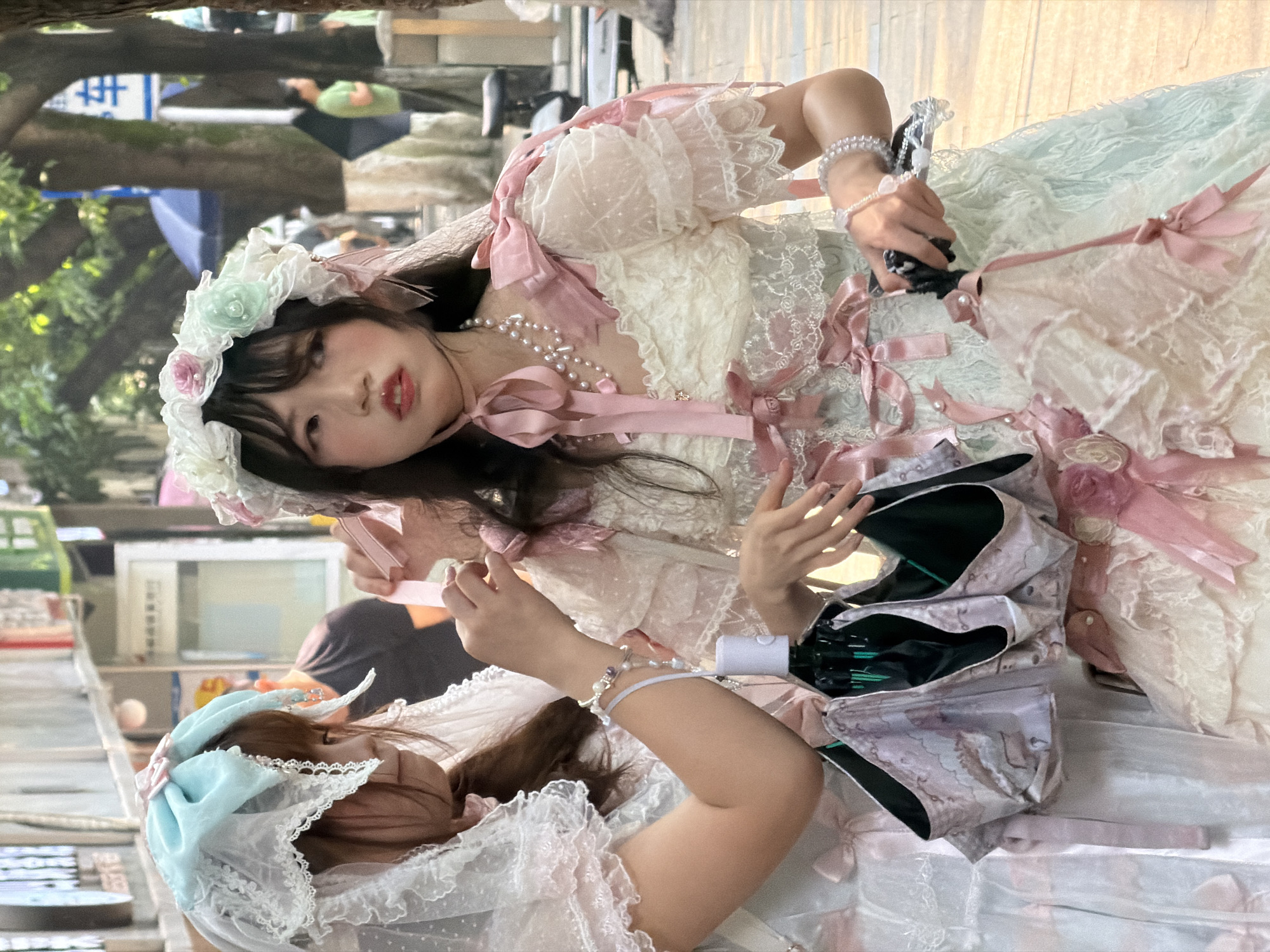 Two young women, dressed in elaborate Lolita fashion, are observed on a paved street in Yuzhong District, China, during daylight hours.
The woman on the left, visible in profile, has long brown hair and wears a white lace bonnet adorned with a light blue bow and a pink accent. Her dress is white lace with pink bows. She wears multiple pearl and beaded bracelets on her right wrist. Her right hand is raised, adjusting a detail on the second woman's attire. Her left hand holds a partially closed umbrella, patterned with light purple/pink designs and a black interior.
The woman on the right, positioned centrally, faces slightly left with a direct gaze. She has dark hair with bangs, pronounced cheek blush, and red lipstick. She wears a floral headpiece composed of white, pink, and light green fabric flowers and ribbons. Her dress is multi-layered, featuring a base of light green and white lace, adorned with numerous pink bows and ruffles on the bodice and sleeves. A multi-strand pearl necklace with a small pendant and a thin pink ribbon choker are visible around her neck. Pink ribbons are tied on her shoulders. Her left hand is near the partially closed umbrella, while her right hand holds a small black object.
The background includes blurry building facades with Chinese signage on the left, and several large tree trunks on the right. A blue and white street sign with Chinese characters, potentially indicating "parking," is partially visible behind one tree. Other blurry figures are discernible in the deeper background. The ground is paved.