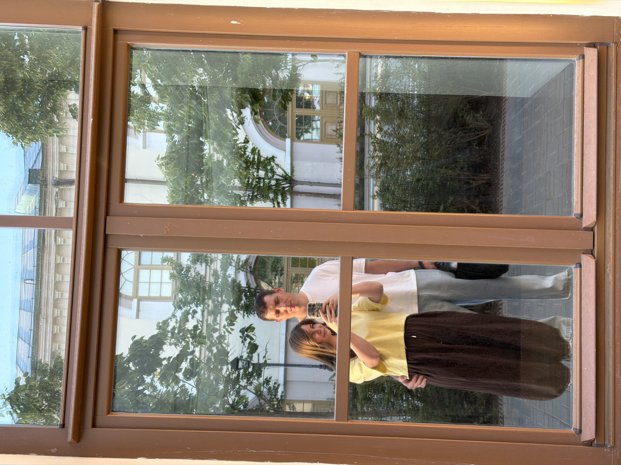 A reflection of a couple is captured within the brown wooden frame of a multi-pane window in Kazan, Russia. The woman, positioned on the left, wears a yellow short-sleeved top and a dark brown long skirt. She holds a smartphone with both hands, aiming it to capture a selfie, and looks at the screen. Her left arm is wrapped around the man's lower back. The man, standing slightly behind and to the right, wears a white short-sleeved t-shirt and light-colored pants. His right arm is placed around the woman's upper back, and he holds a black bag in his left hand, looking straight ahead.
The window panes reflect an outdoor urban environment behind the couple. The upper panes show a clear blue sky, tree foliage, and the upper section of a building featuring a distinctive blue patterned roof and a light-colored classical facade with architectural details. The lower panes reflect numerous green trees, an ornate black street lamp positioned behind the woman's head, and a large light-colored building with multiple arched windows and a prominent arched wooden double door. The ground surface reflected below consists of rectangular paving stones.