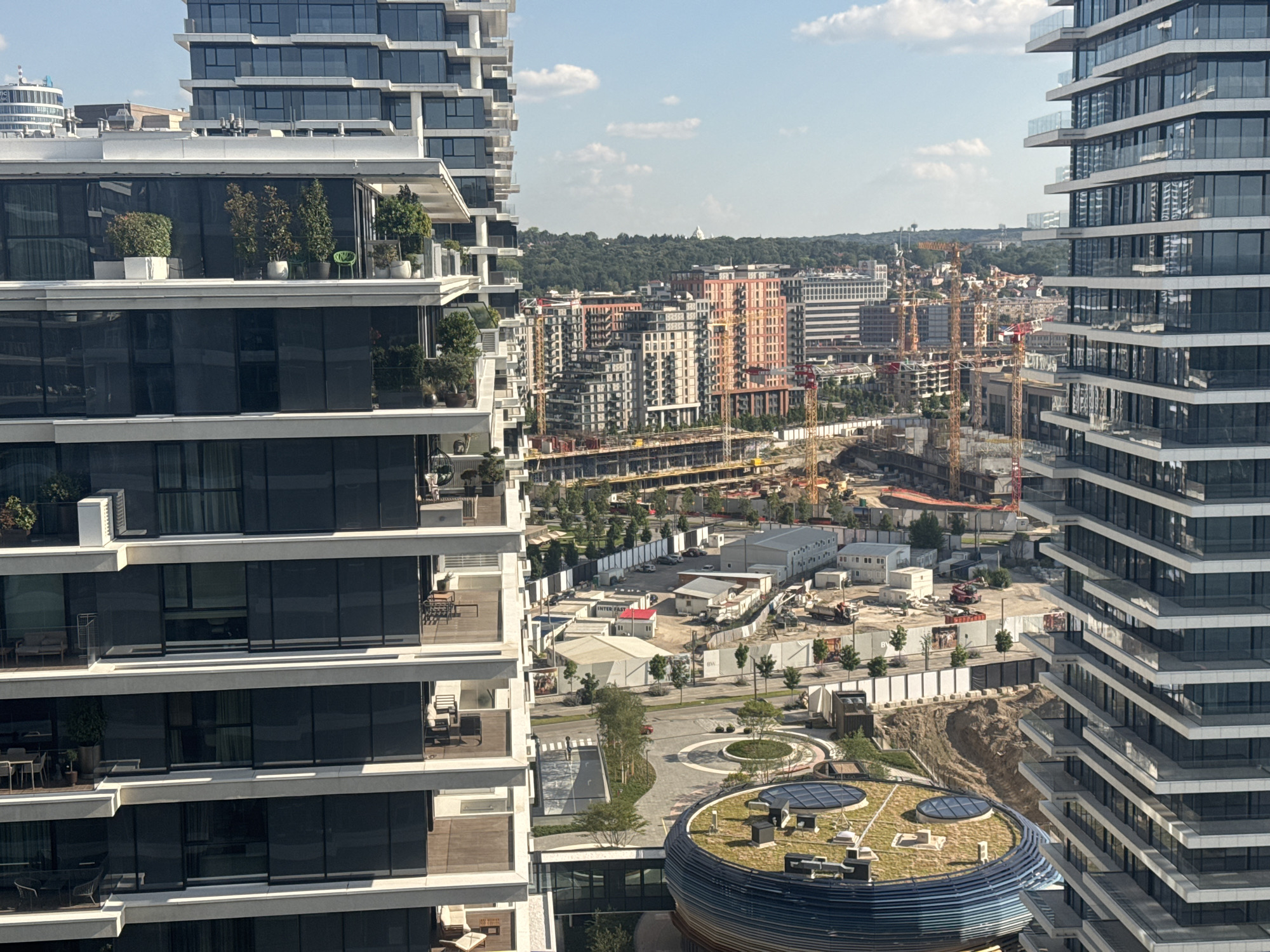 The image captures an aerial view of a construction site in Belgrade, Serbia, alongside completed modern residential buildings. In the foreground, two tall, multi-story residential buildings with balconies and large glass windows are prominently featured. To the left, the balconies have planters with greenery and outdoor furniture. To the right, the building's facade displays stacked horizontal elements.
Beyond these structures, the scene transitions to an active construction zone. Several cranes are visible, indicating ongoing development. Various buildings in different stages of construction are present, some with exposed frameworks and others with completed facades. Temporary structures and equipment associated with construction activities are scattered across the site.
In the background, the cityscape of Belgrade unfolds, characterized by a mix of residential and commercial buildings. A line of trees forms a natural backdrop, with a faint white dome-like structure visible on a distant hill, possibly a landmark or monument. The sky is clear with scattered white clouds, suggesting a sunny day. The overall impression is one of urban development and modernization in Belgrade.