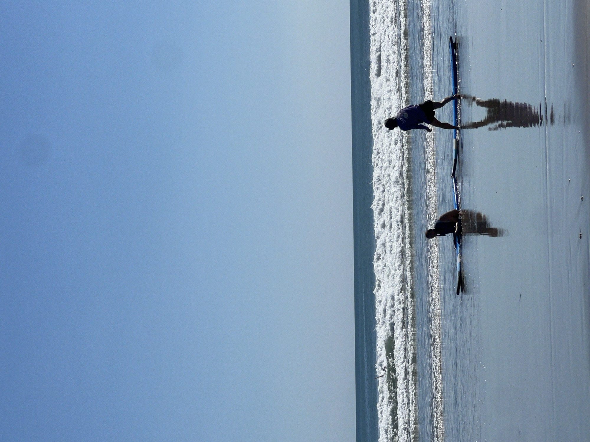 Two individuals are on surfboards in shallow water at Kuta Beach, Bali, Indonesia. The person on the left is kneeling on a blue and black surfboard, facing away from the camera. The person on the right is standing on a blue surfboard, facing towards the ocean and away from the camera. They are wearing a purple long-sleeved shirt and dark shorts. White-capped waves are breaking in the background. The sky is clear and blue. The wet sand reflects the figures and the sky.